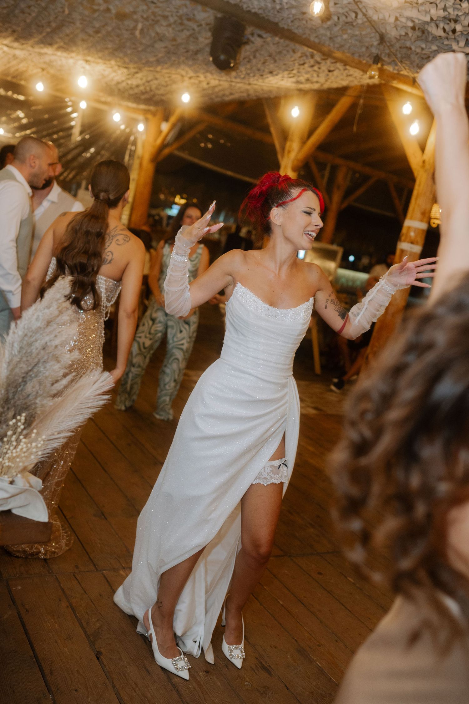 People dancing and celebrating at a rustic wedding reception with string lights overhead on a wooden floor.