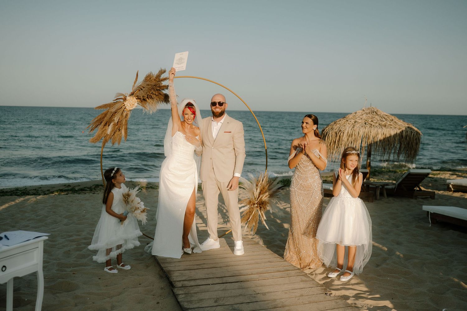 A group in white attire walks along a wooden pier at sunset by the ocean.