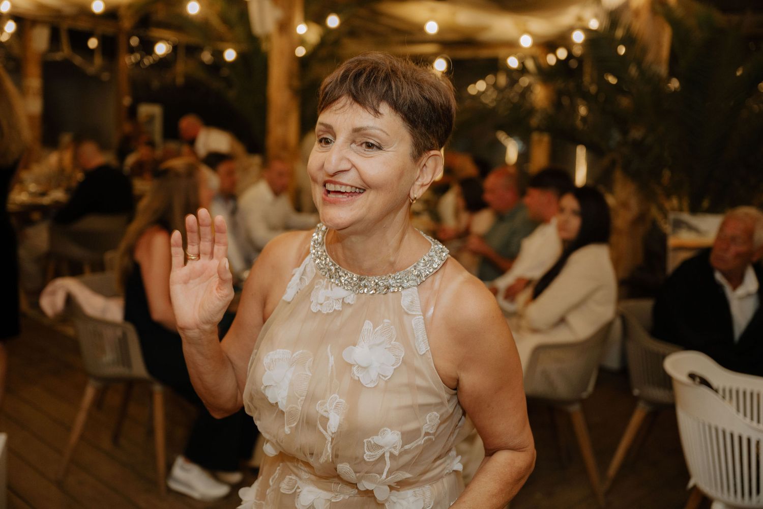 A person in a white lace dress dances joyfully at a wedding reception with string lights in the background.