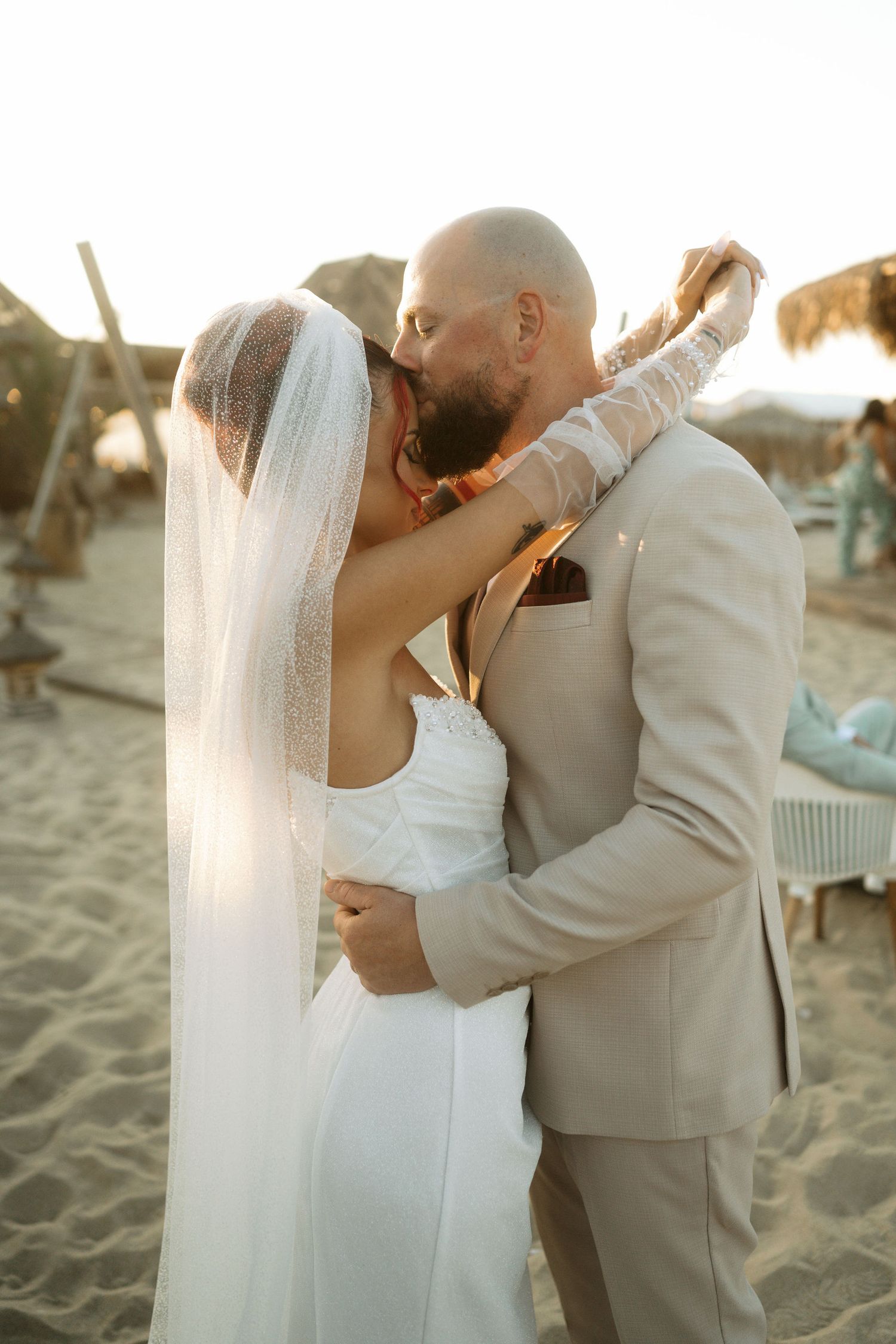 A couple shares a romantic kiss on the beach during sunset in their wedding attire.