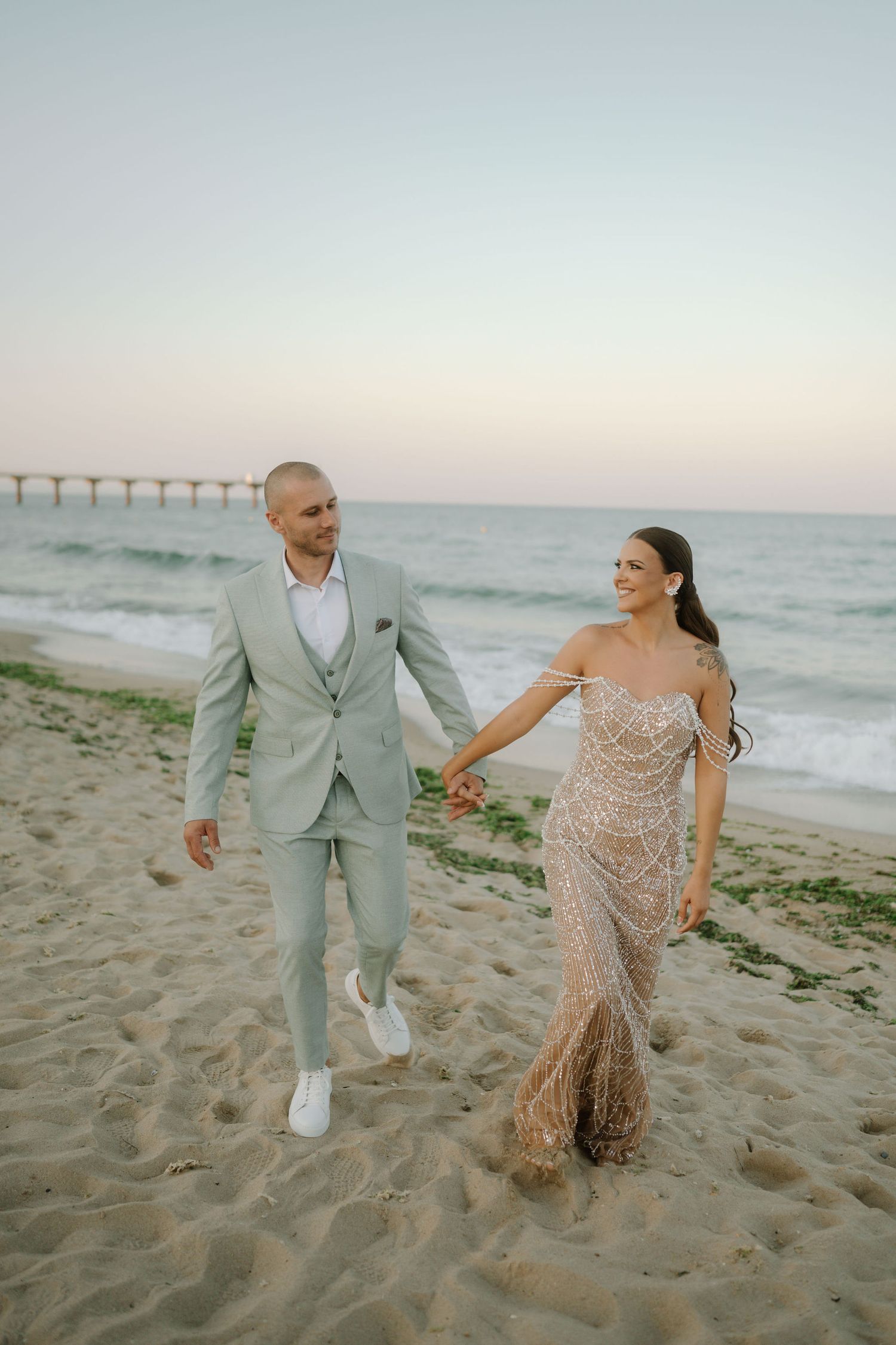A couple in formal attire walks joyfully along a sandy beach at sunset with waves in the background.