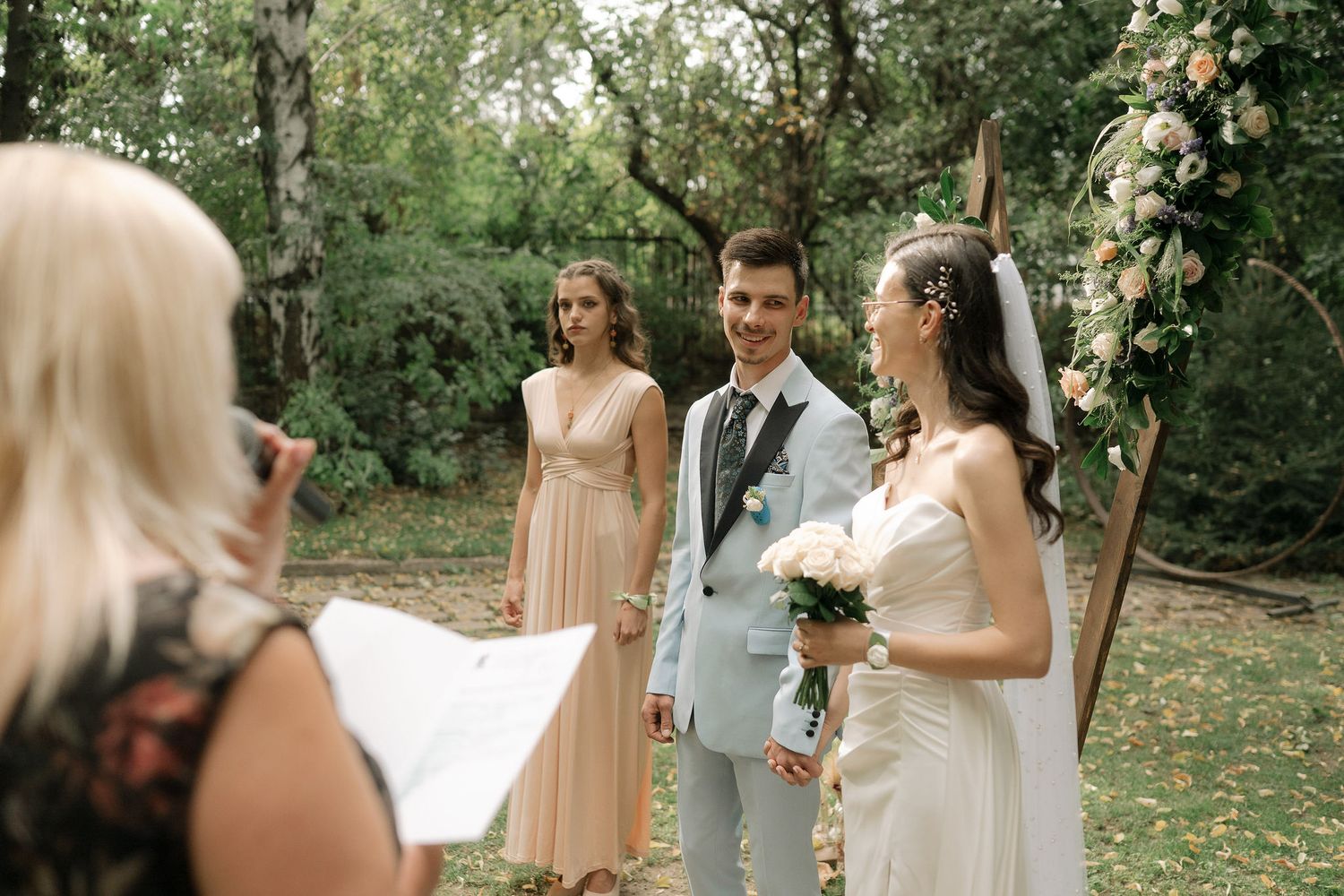 A wedding party celebrates outdoors in a natural garden setting with soft peach bridesmaid dresses and floral decor.