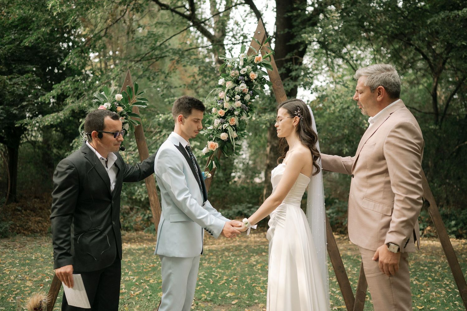 An outdoor wedding ceremony takes place in a lush garden setting with an officiant and guests gathered around the couple.