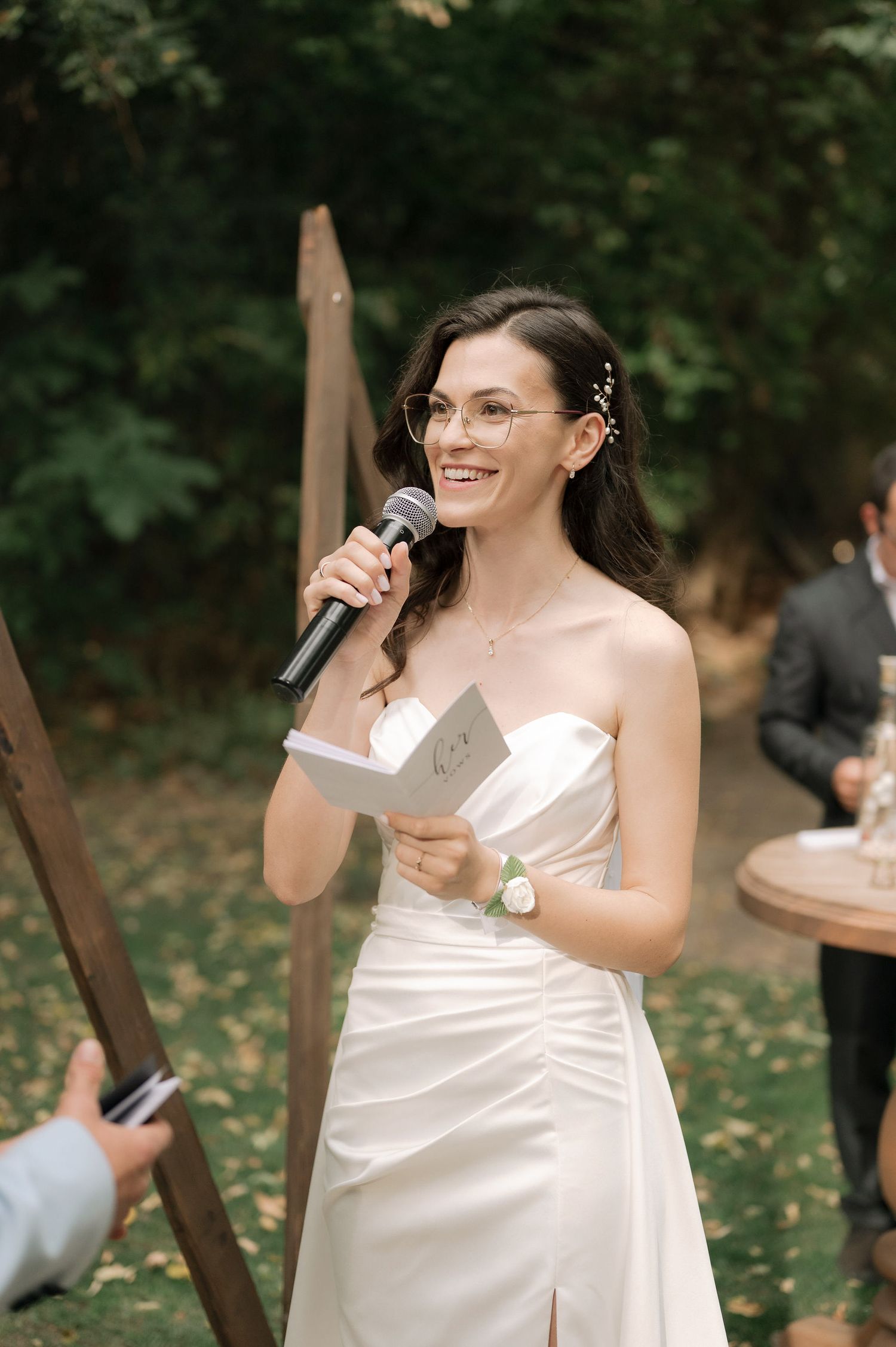 A person in a white dress holding a microphone and speaking at an outdoor event in a garden setting.