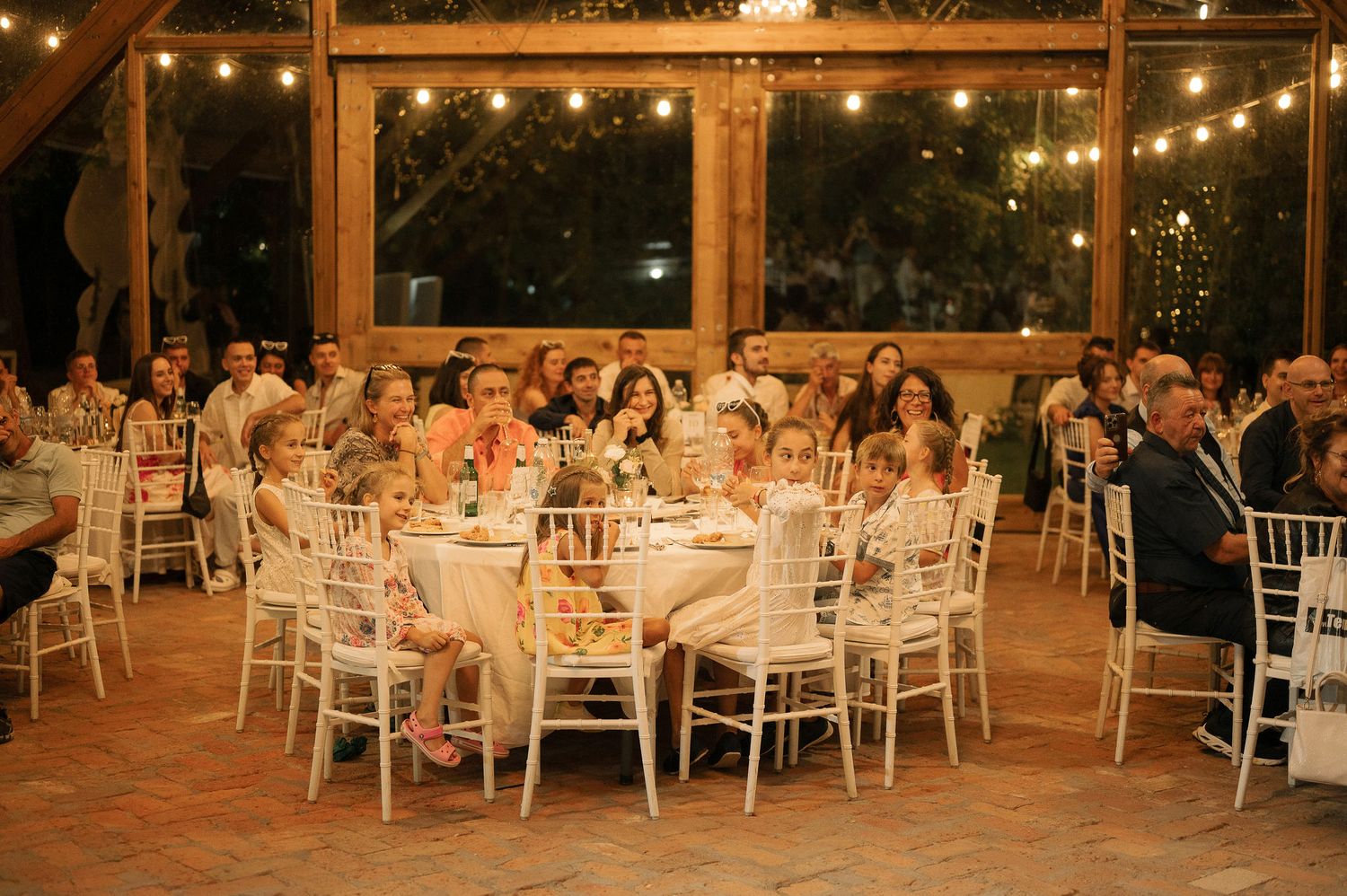 Elegant outdoor wedding reception under string lights with white chiavari chairs and round dining tables.