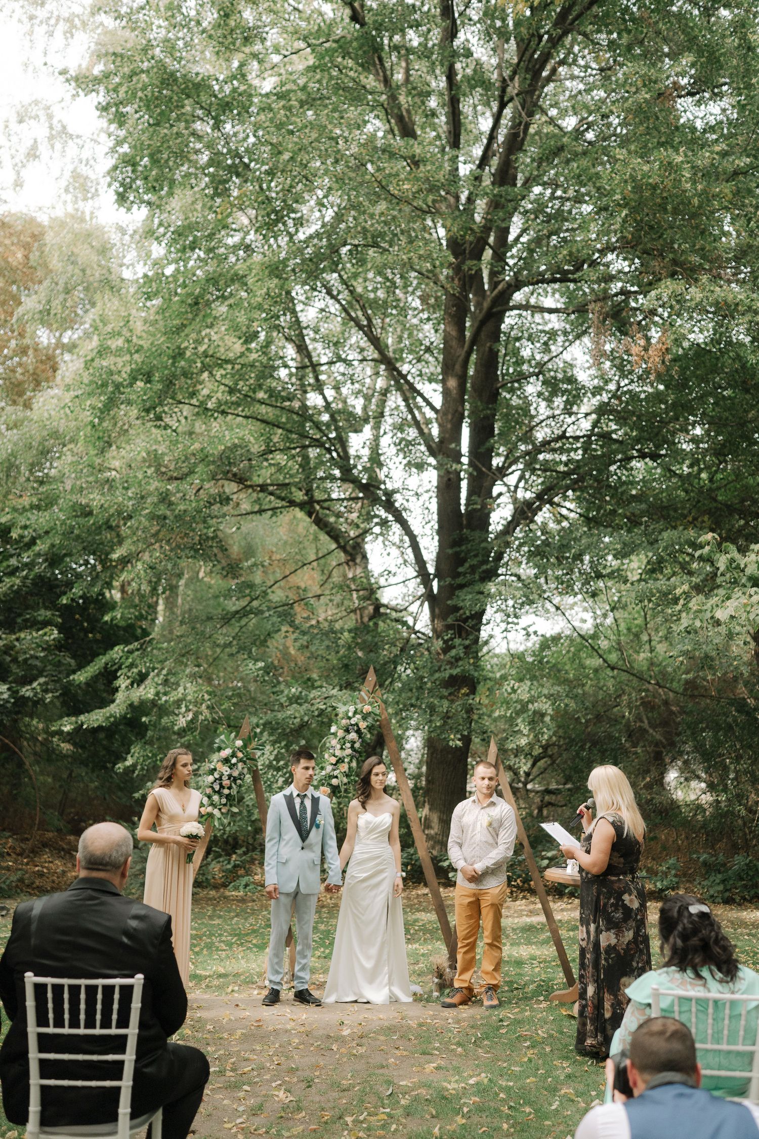 Wedding ceremony taking place outdoors in a wooded setting with guests seated on white chairs among leafy trees.