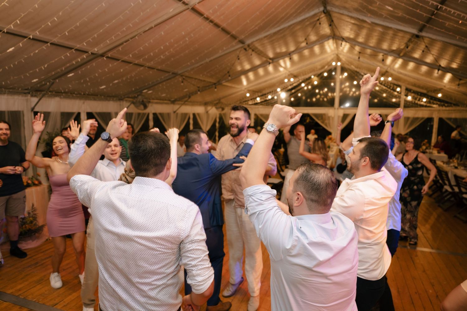 Wedding guests dance in a line formation under twinkling string lights at an indoor reception venue.