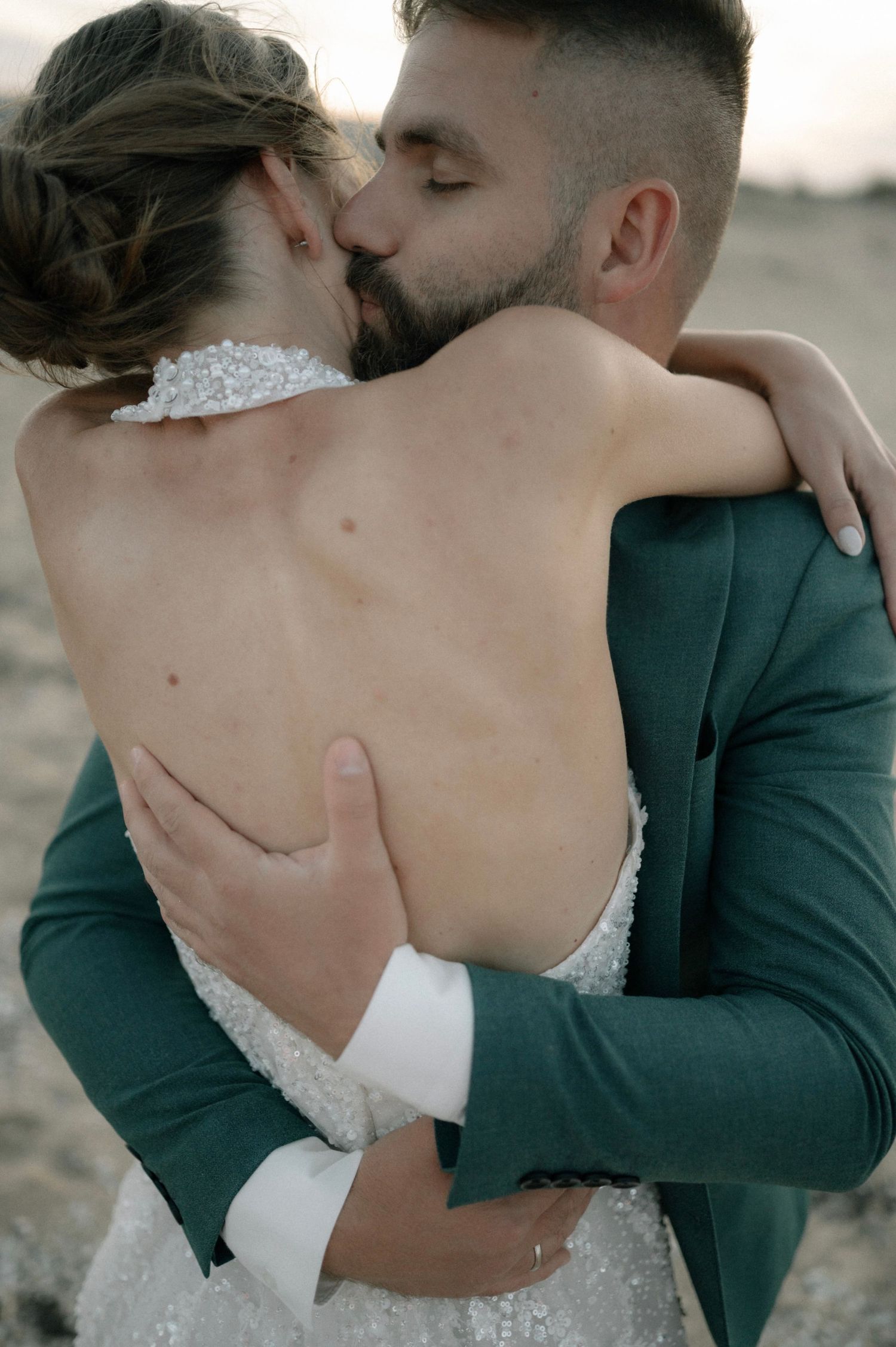 A couple shares a romantic kiss, the bride in a backless white gown embraced by the groom in a green suit outdoors.