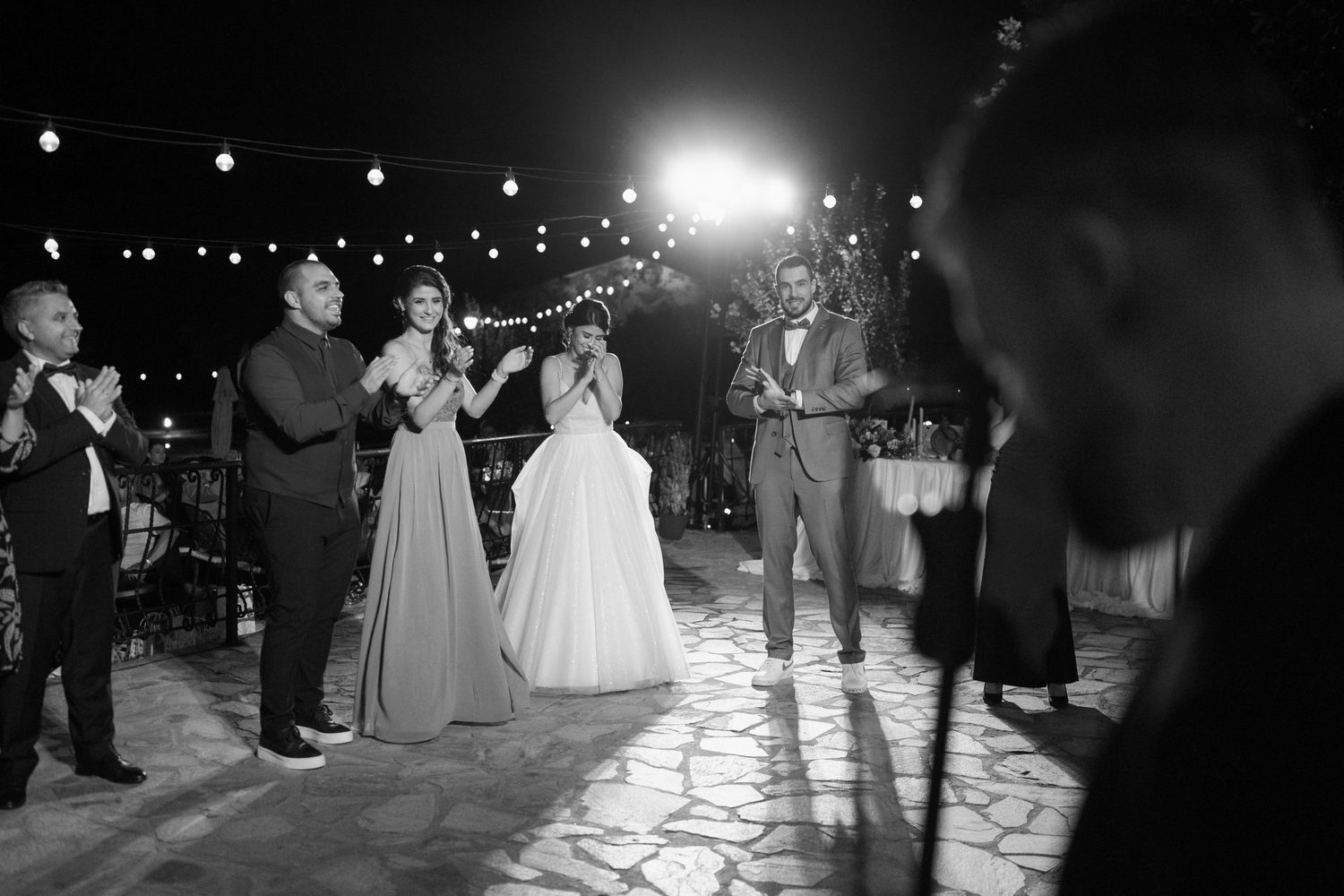 Black and white photo of a wedding reception dance floor with string lights overhead and guests celebrating at night.