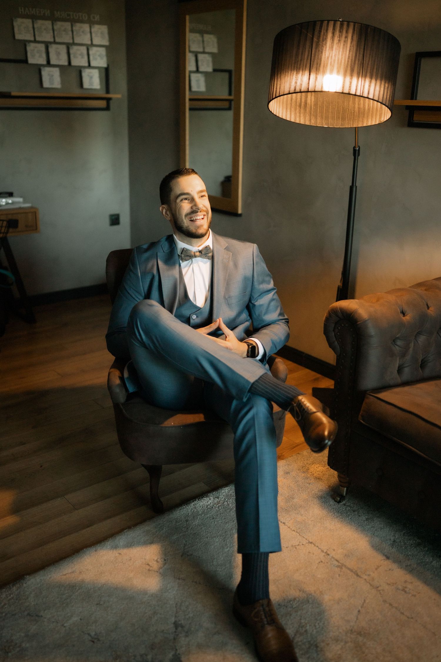 A well-dressed person in a blue suit and bowtie sits relaxed in a leather armchair next to a floor lamp in a cozy room.