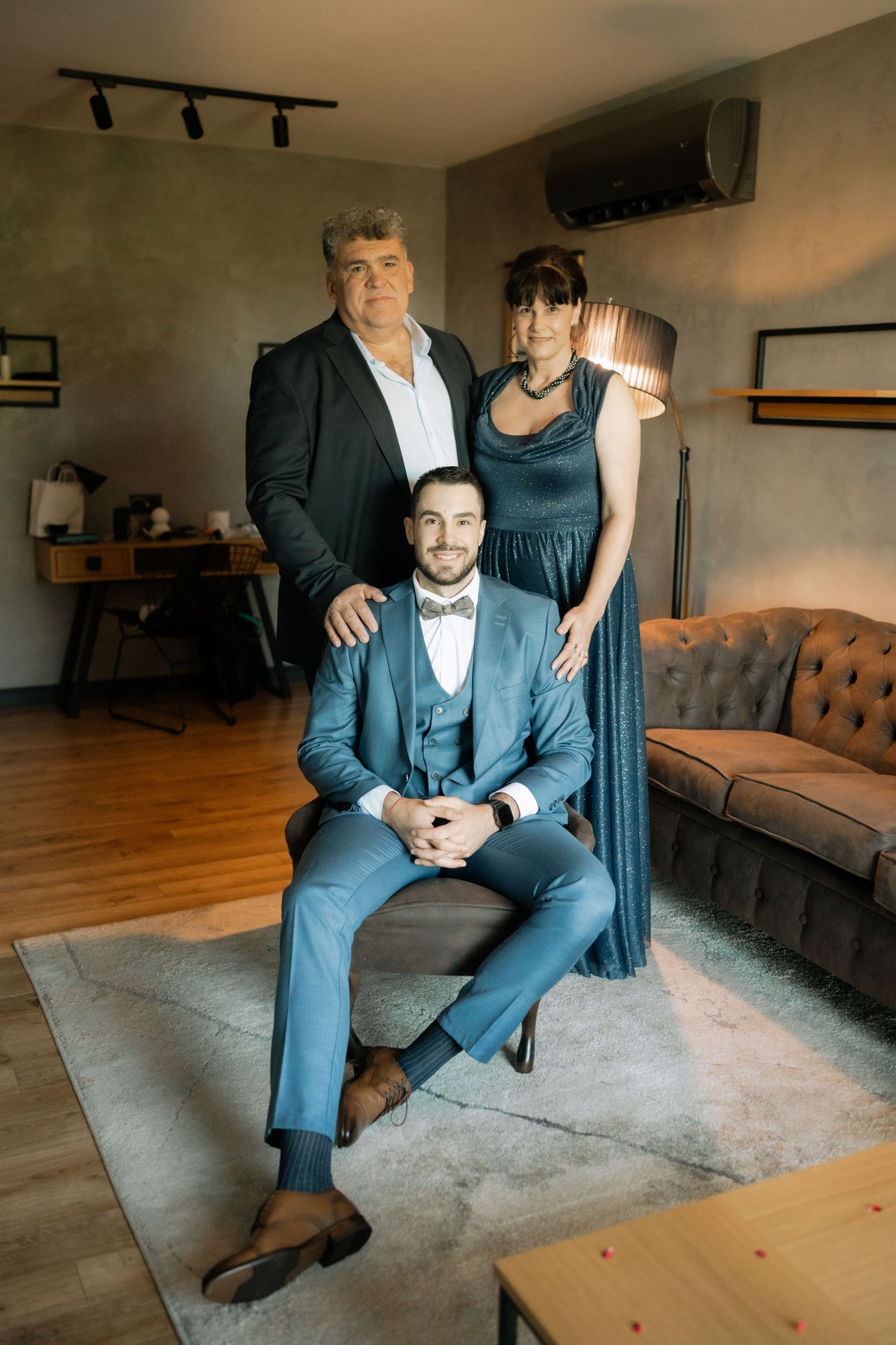 Three people in formal attire pose together in a modern living room with hardwood floors and track lighting.