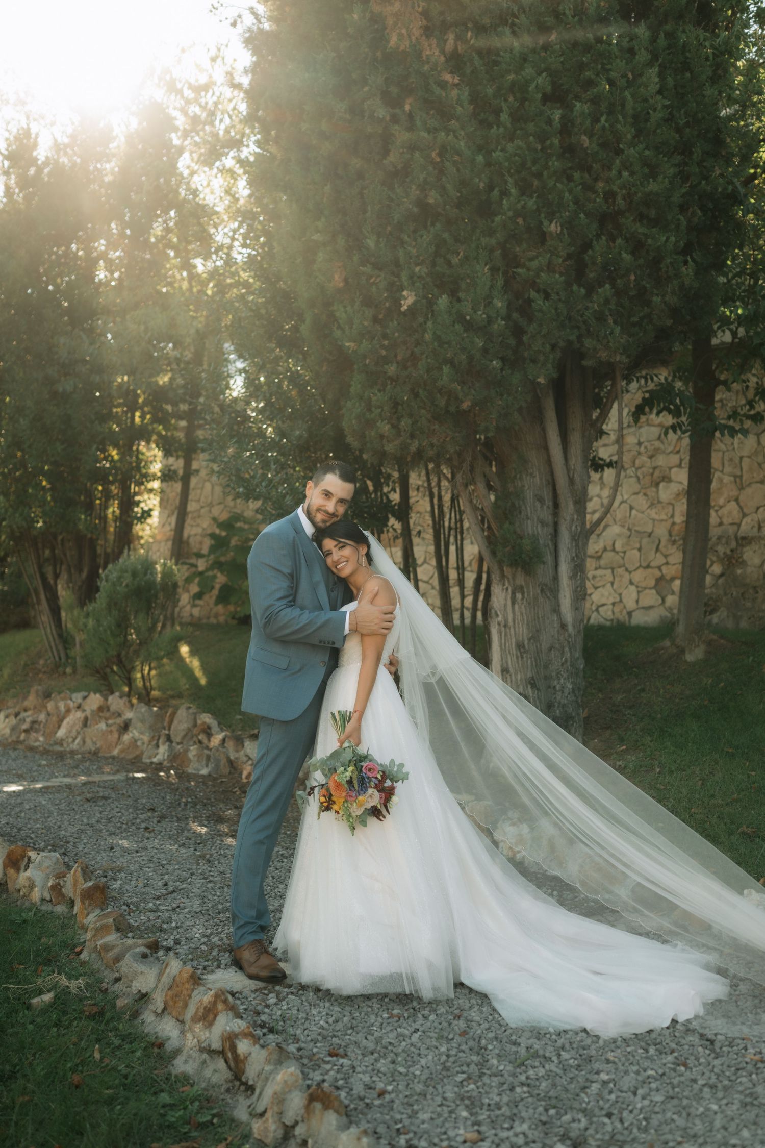 A romantic wedding couple embraces on a gravel path at sunset, with trees and stone walls in the background.