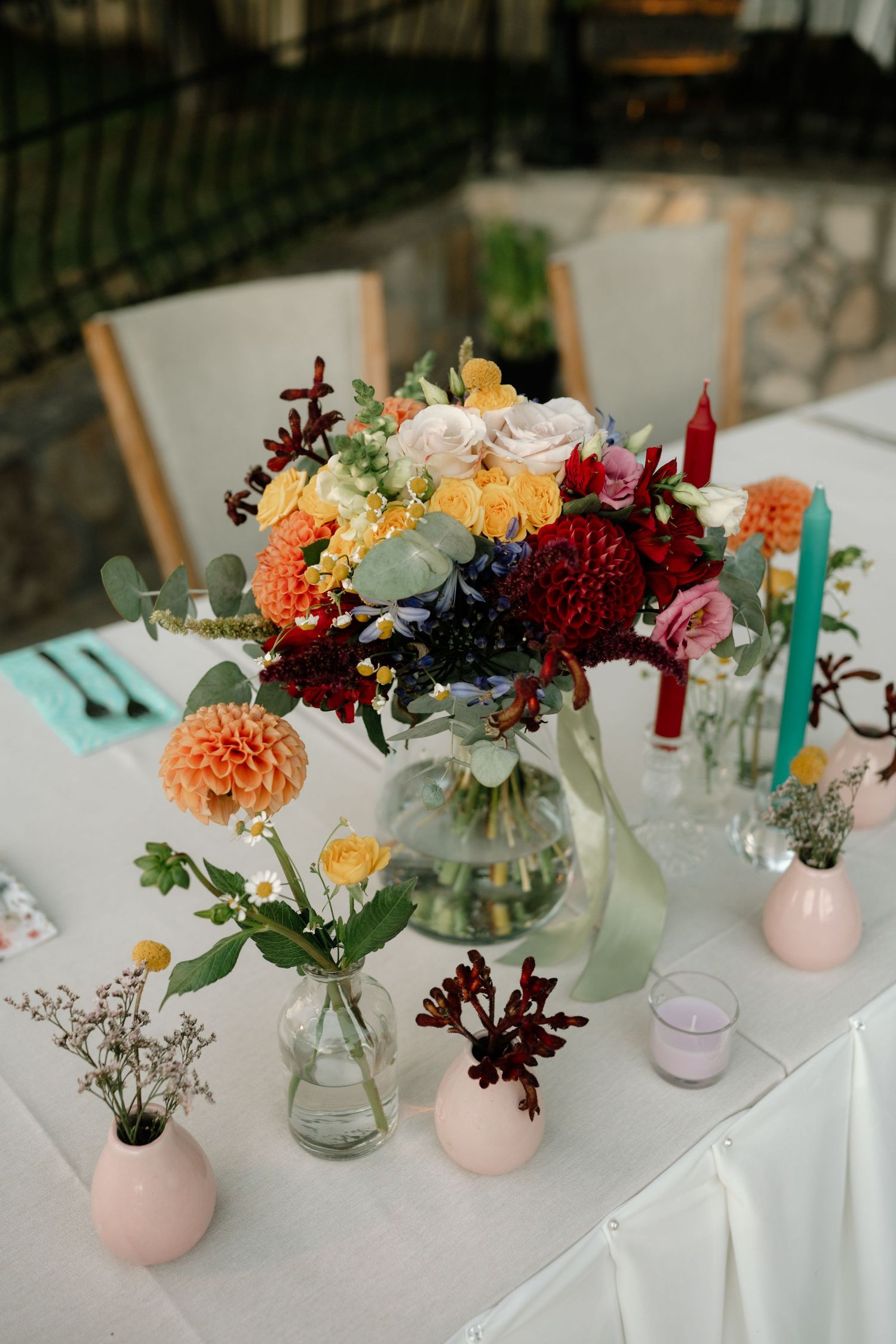 Colorful floral centerpieces with dahlias and mixed flowers in glass vases and pink holders arranged on white tablecloths.