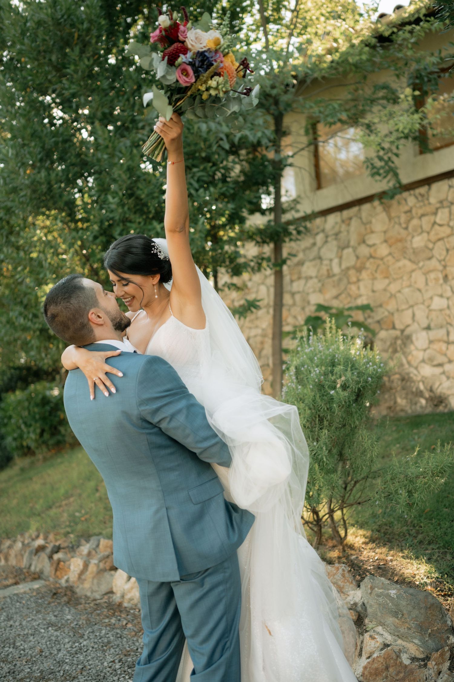 Romantic wedding couple dancing together in a garden setting with stone walls and lush greenery at sunset.
