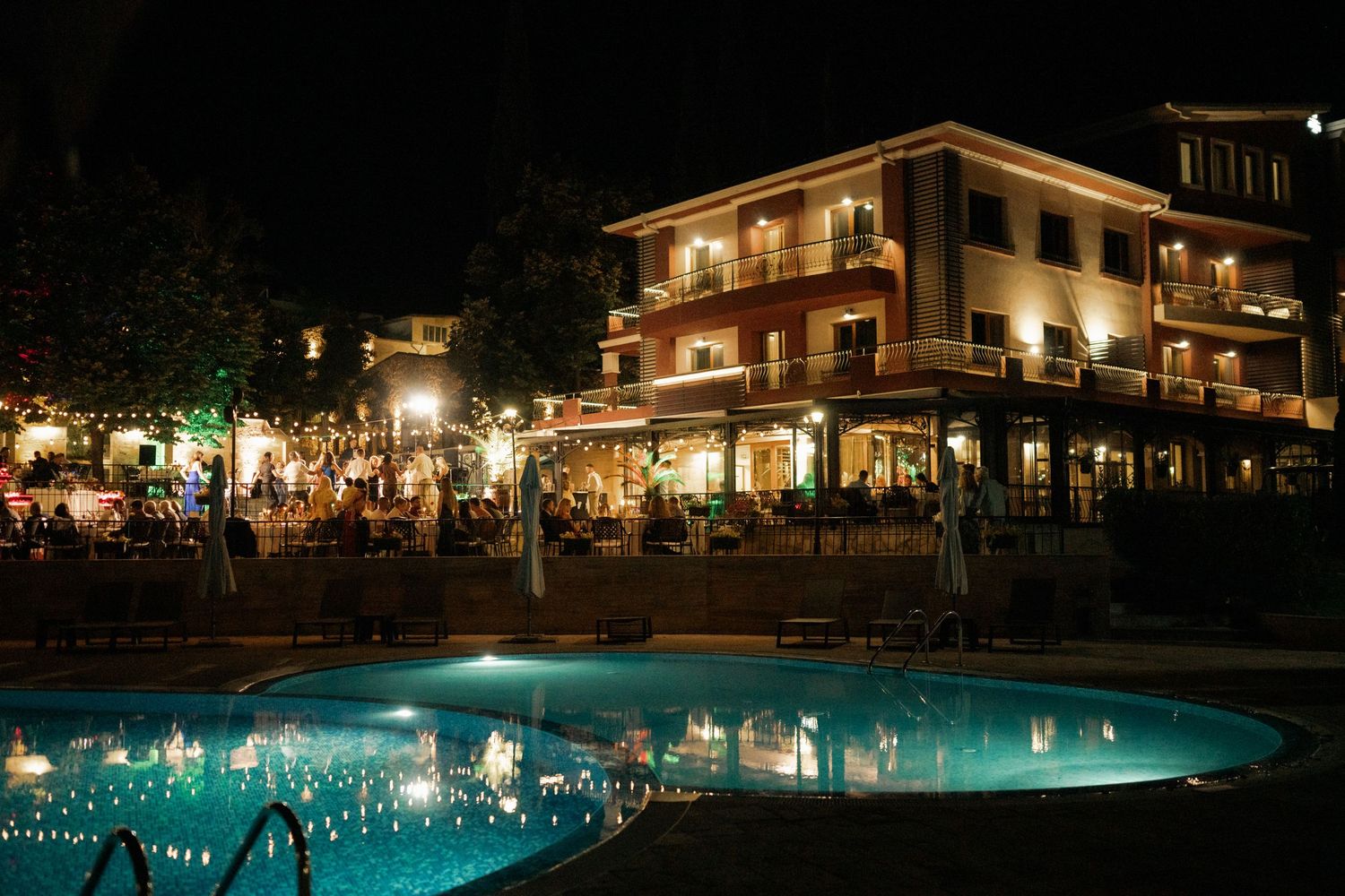A luxurious hotel with illuminated swimming pool at night showing guests dining on the terrace and outdoor seating areas.