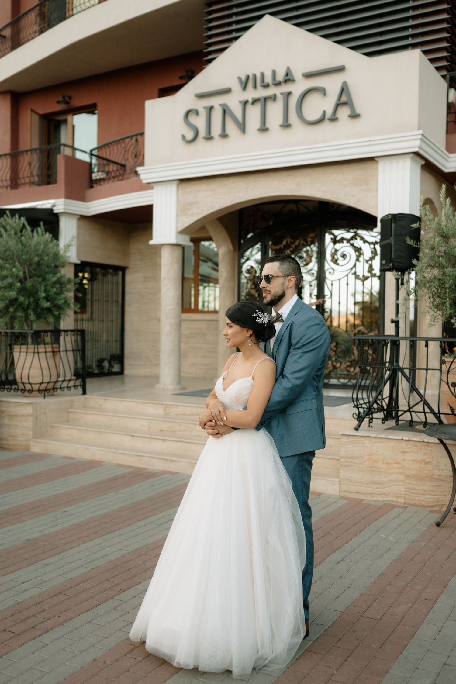 A couple in wedding attire stands embracing outside Villa Sintica, a venue with elegant stone architecture and iron gates.