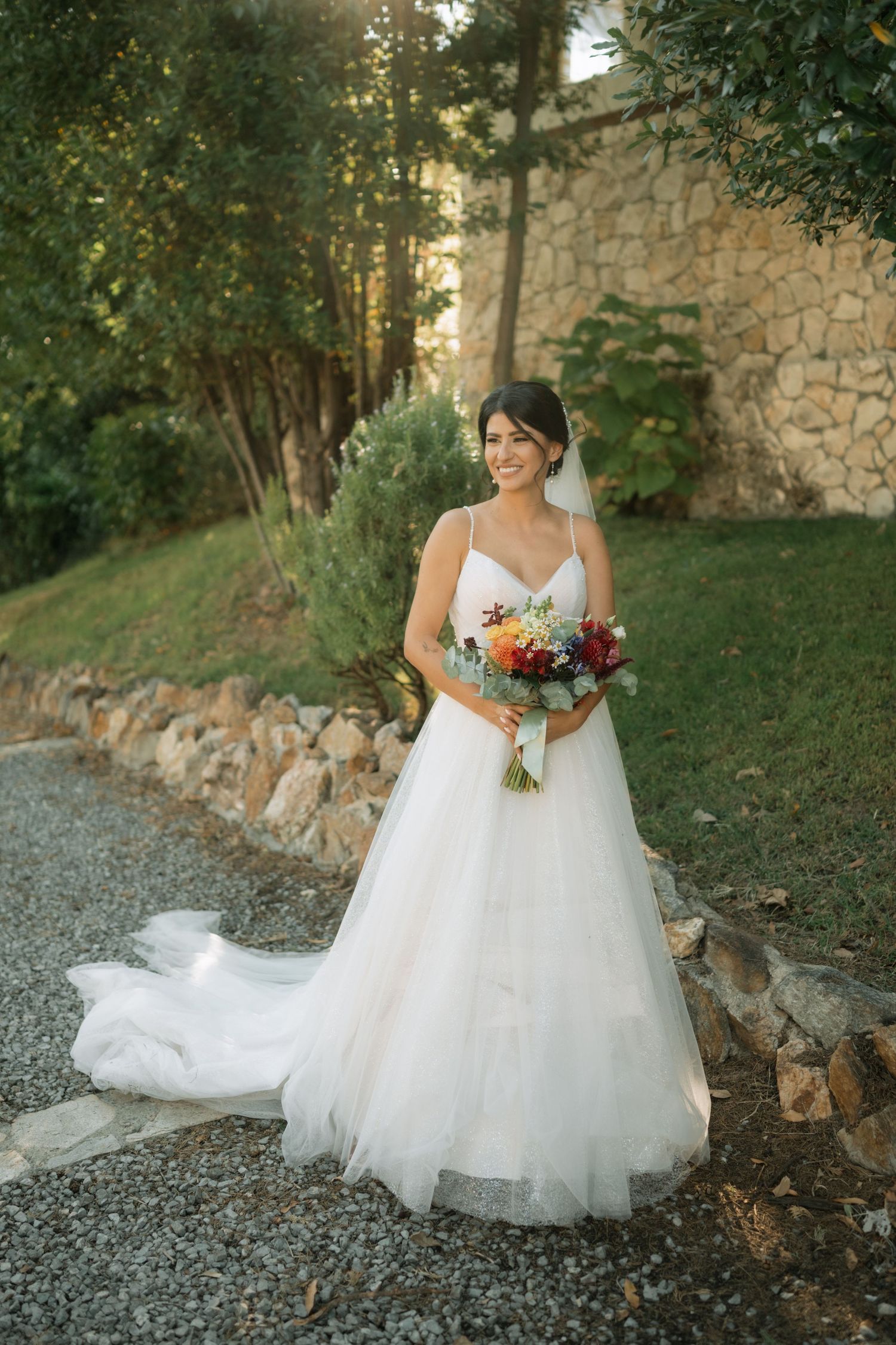 A bride in a flowing white wedding dress holds a vibrant autumn bouquet while posing on a gravel path near stone walls.