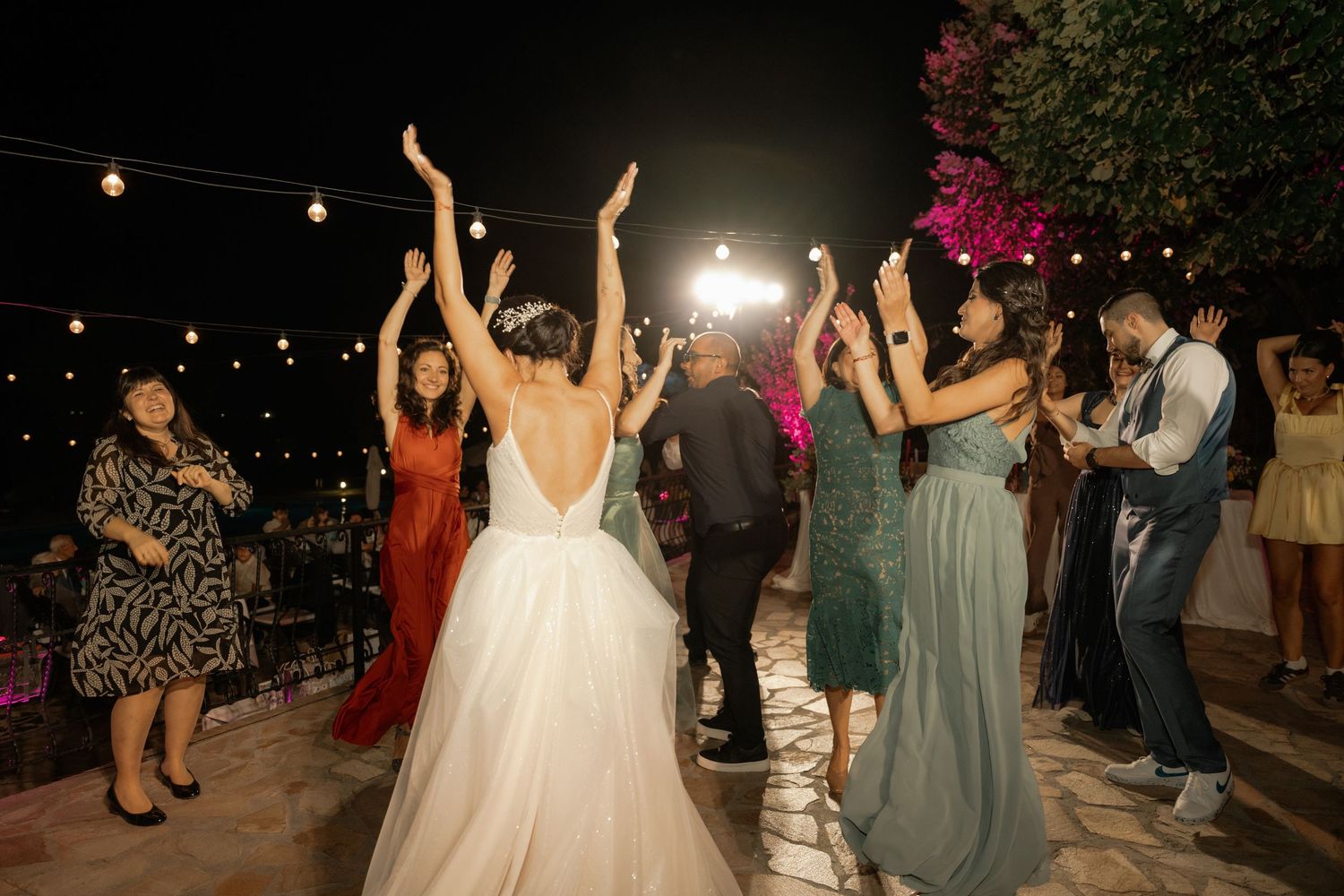 People dancing and celebrating at a lively outdoor wedding reception under string lights at night.