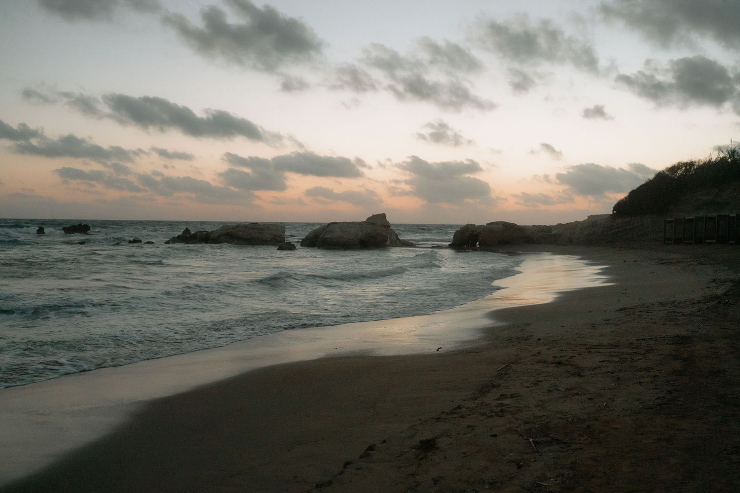 Serene beach at sunset with rocky outcrops, gentle waves, and a cloudy sky reflecting warm hues over the sandy shore.