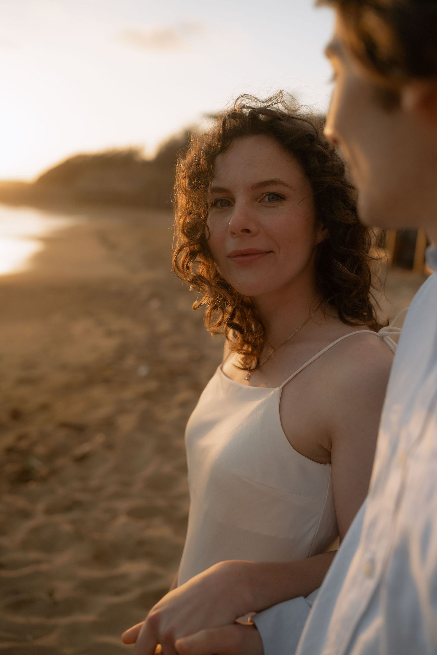 A woman with curly hair in a white sundress smiles softly at sunset on a sandy beach, holding hands with a companion.