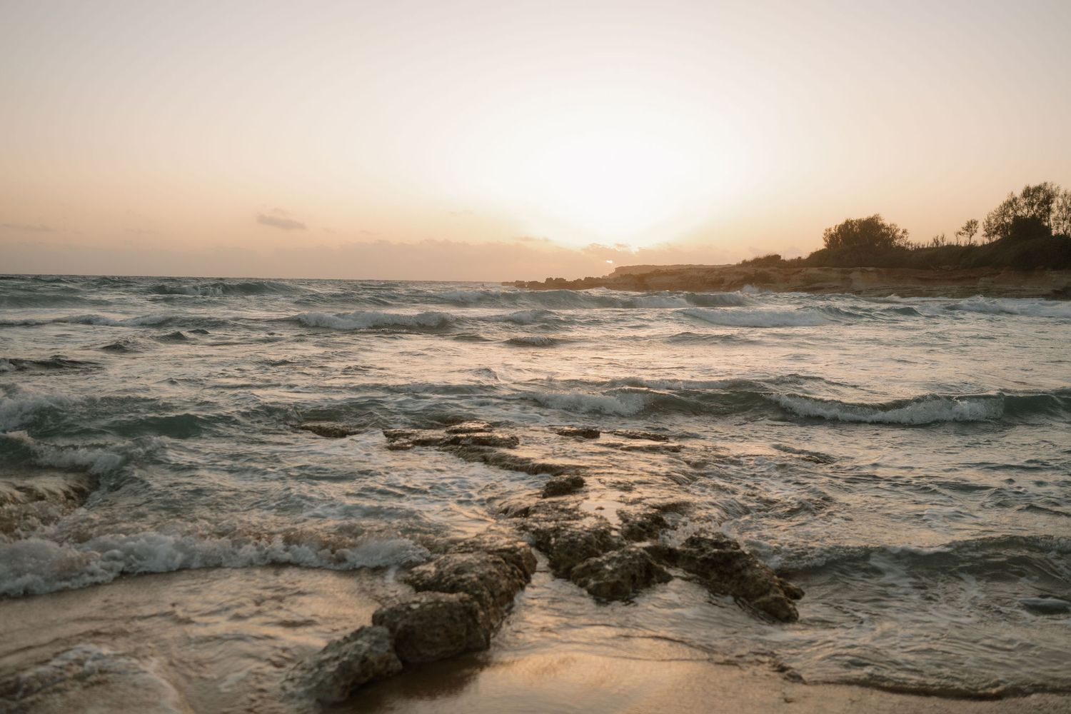 Rocky shoreline with ocean waves and golden sunset sky over tropical coastline.