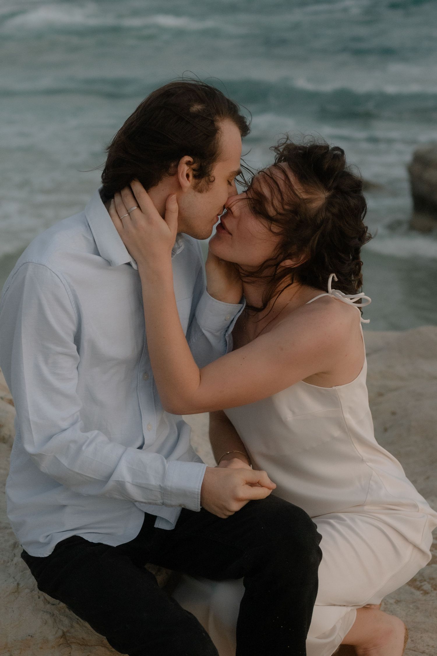 Romantic couple sharing a tender kiss on rocky beach shore with ocean waves in background.