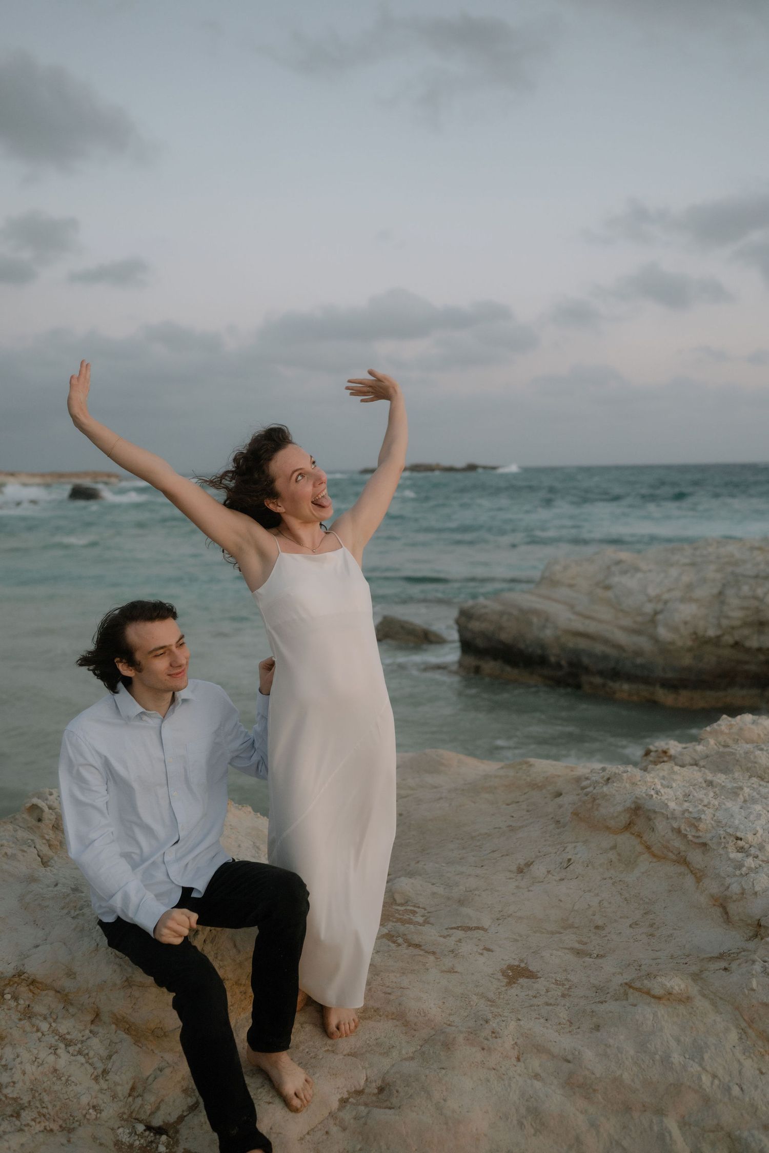 Joyful couple on rocky beach at dusk, woman in white dress with arms raised, man in blue shirt smiling beside her.