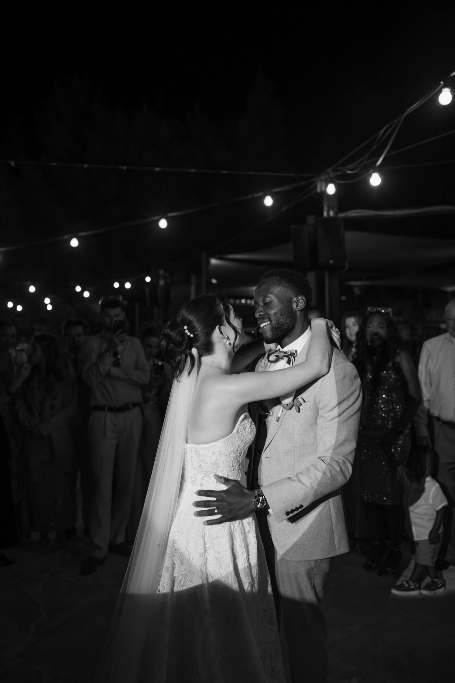 Black and white photo of a bride and groom sharing their first dance under string lights at an outdoor wedding reception.