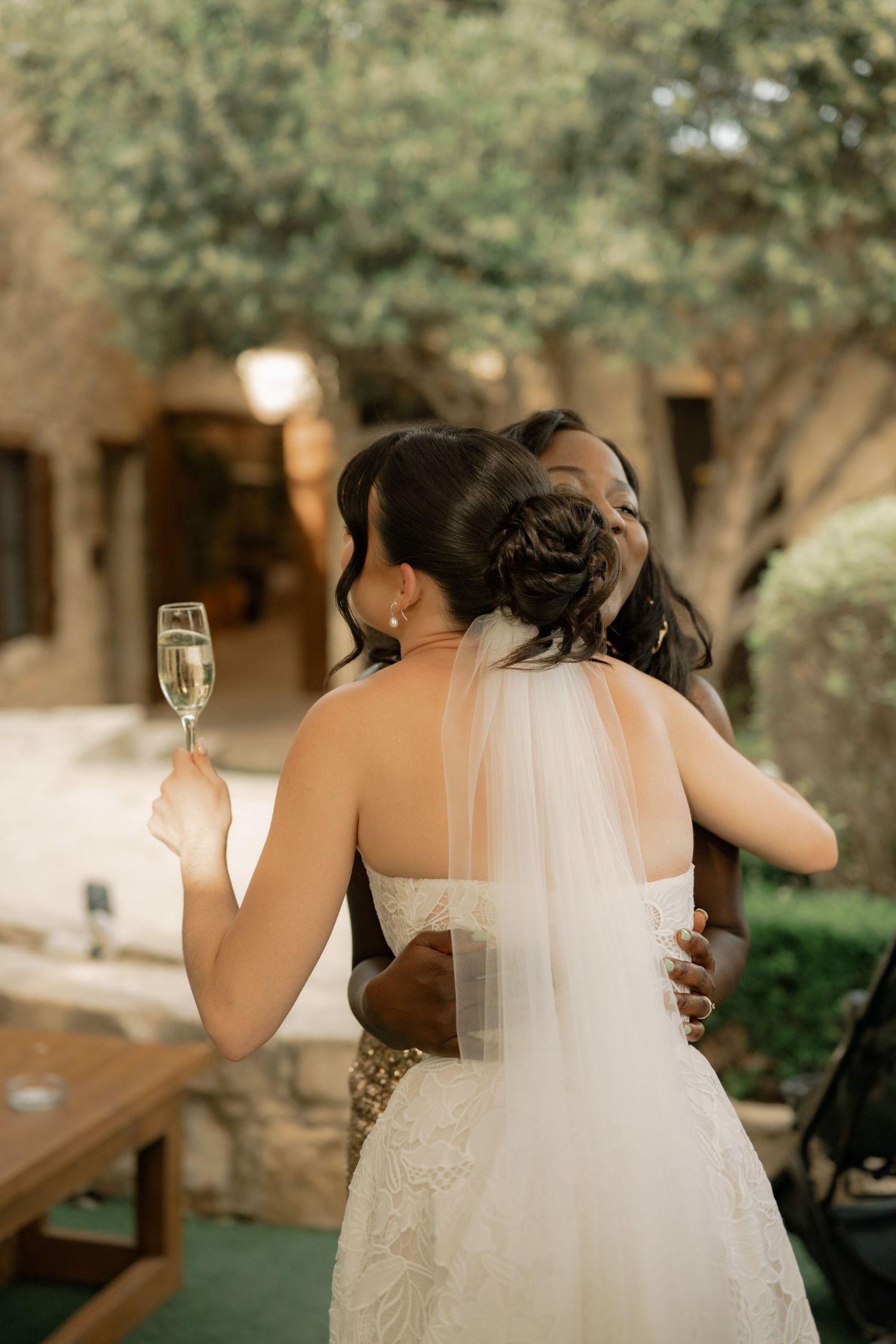 Bride in white gown and veil holds champagne while embracing a guest outdoors at a wedding reception.