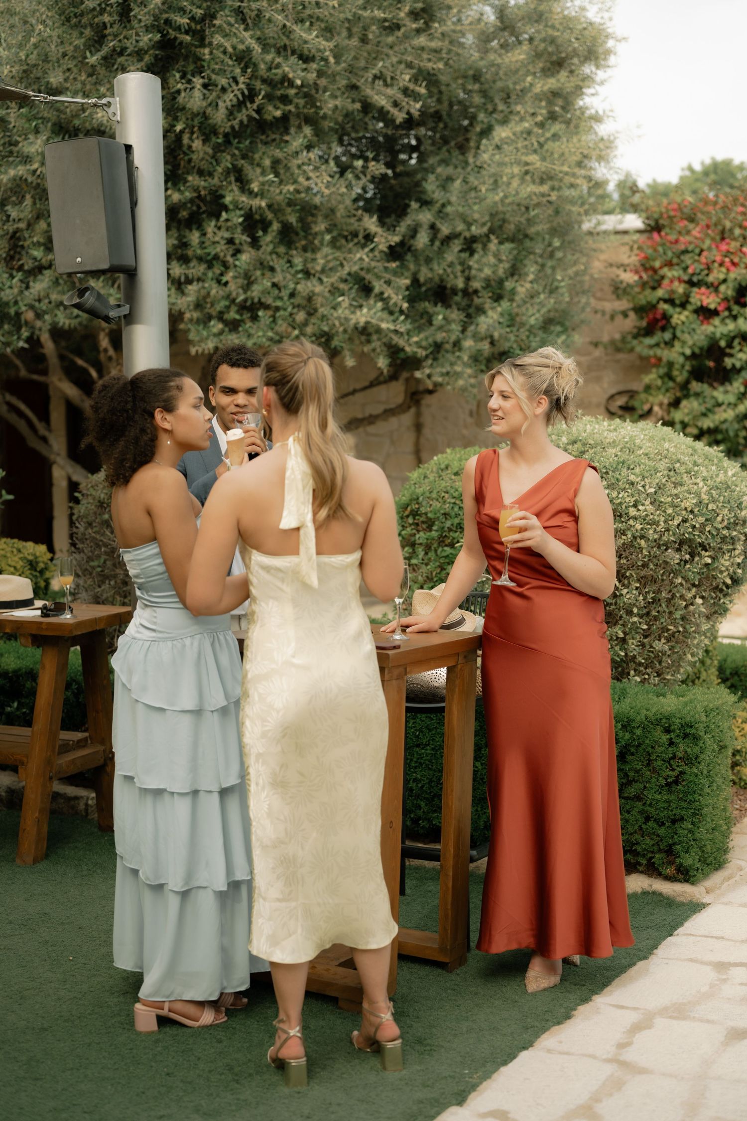 Guests in elegant dresses socializing outdoors at a garden wedding reception with drinks.