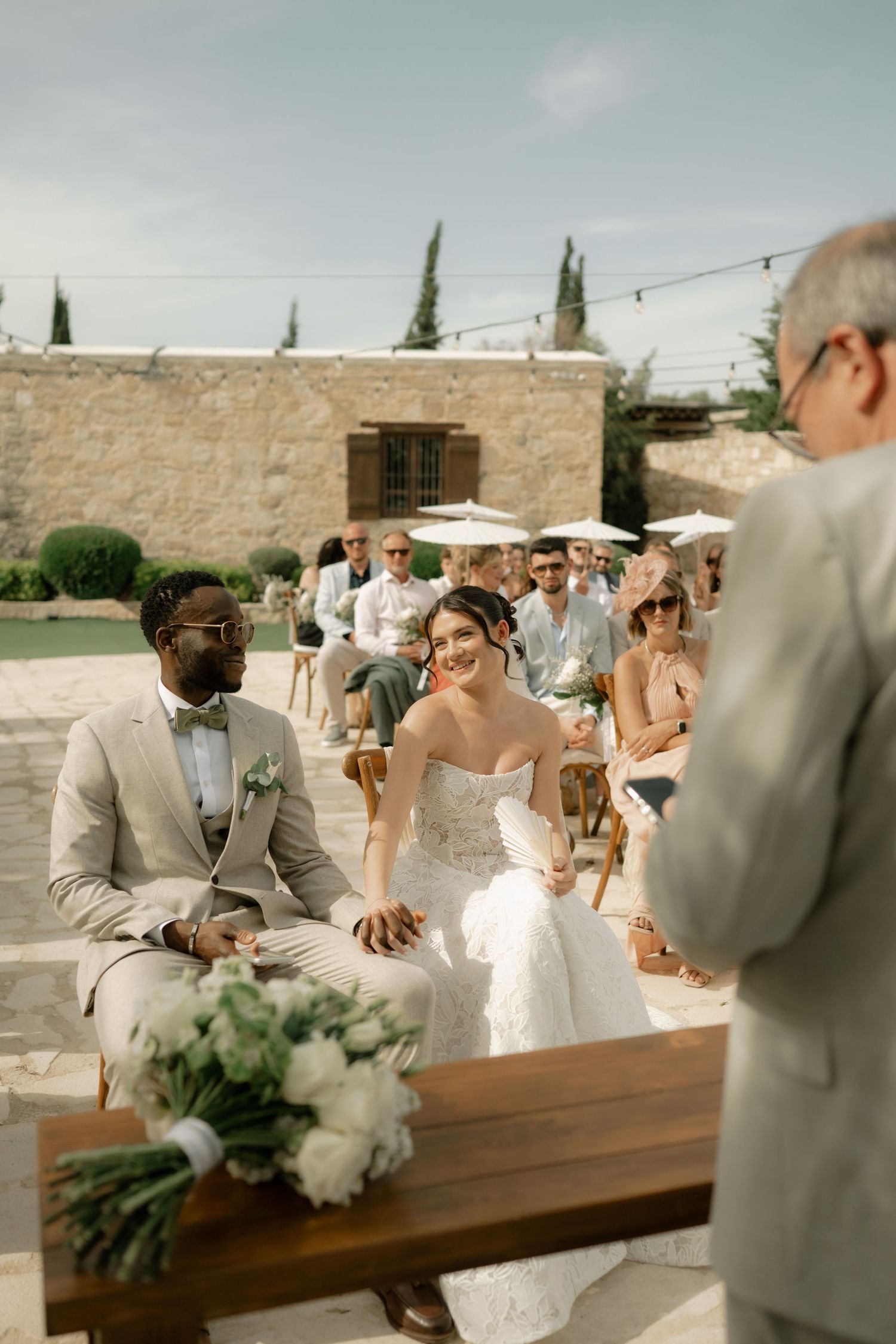 Bride and groom seated at outdoor wedding ceremony with guests behind them at a rustic stone venue.