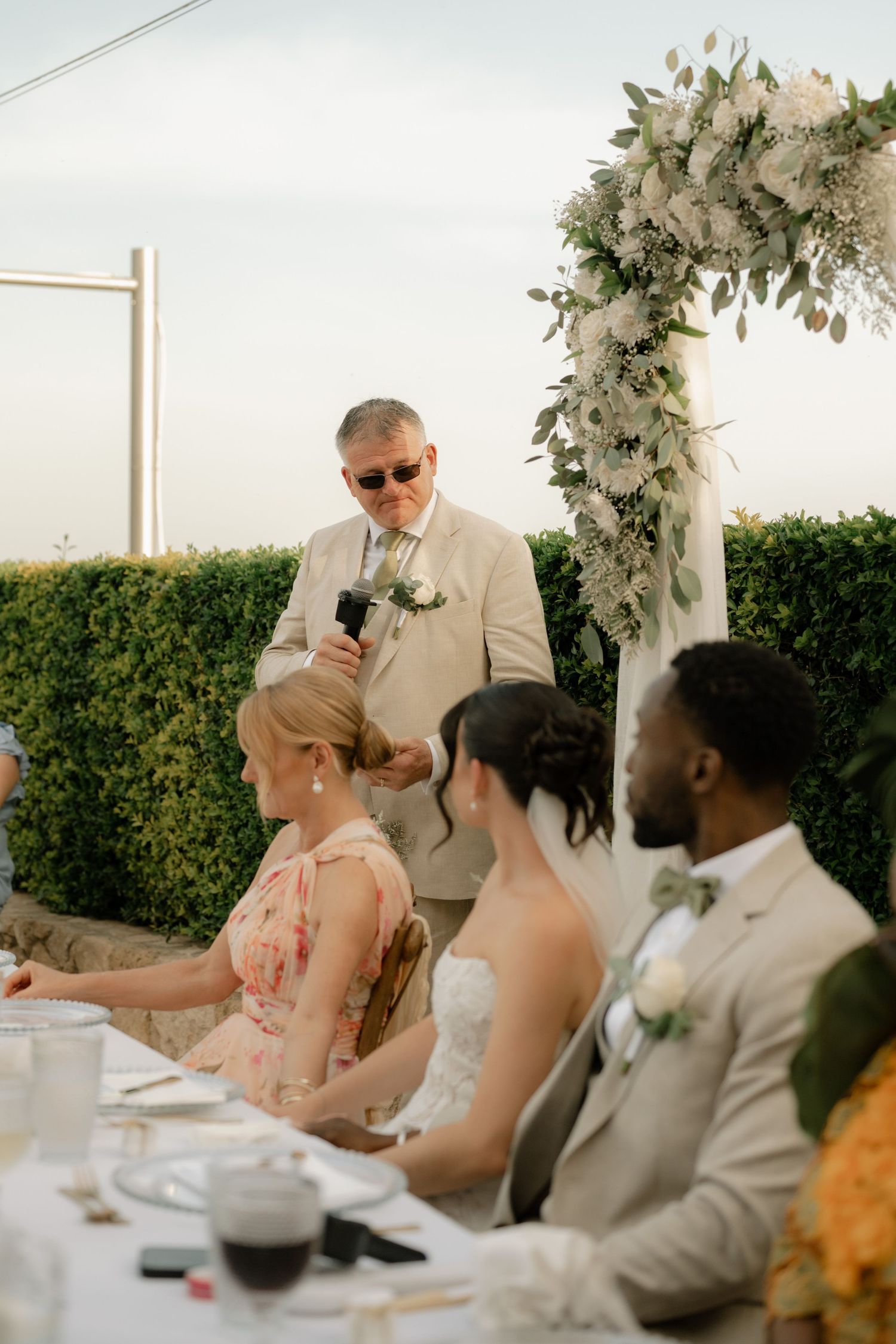 Man in beige suit giving wedding toast at outdoor reception while bride, groom, and guests listen at decorated table.