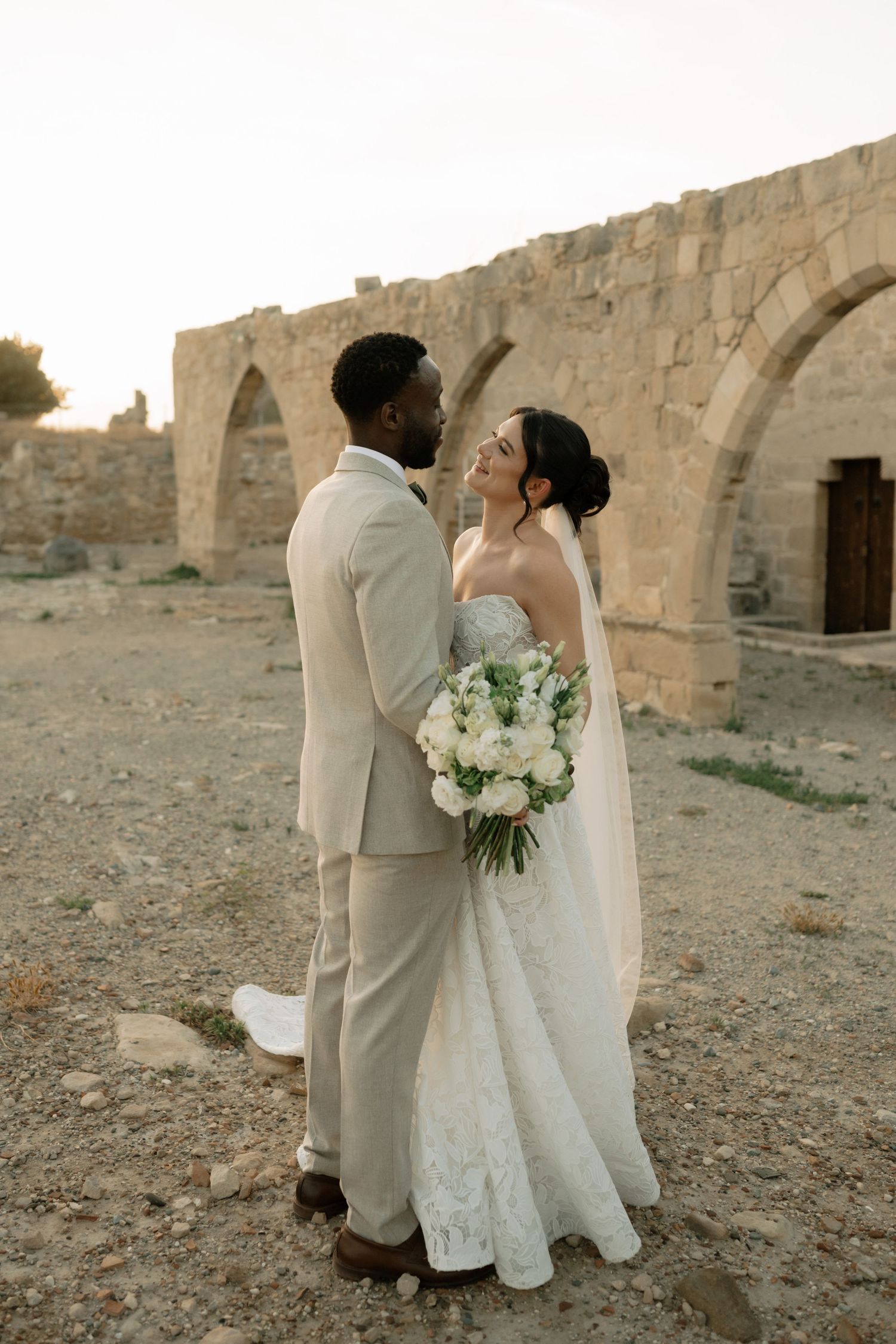 Interracial couple in wedding attire pose before ancient stone arches at sunset, bride holding white bouquet.