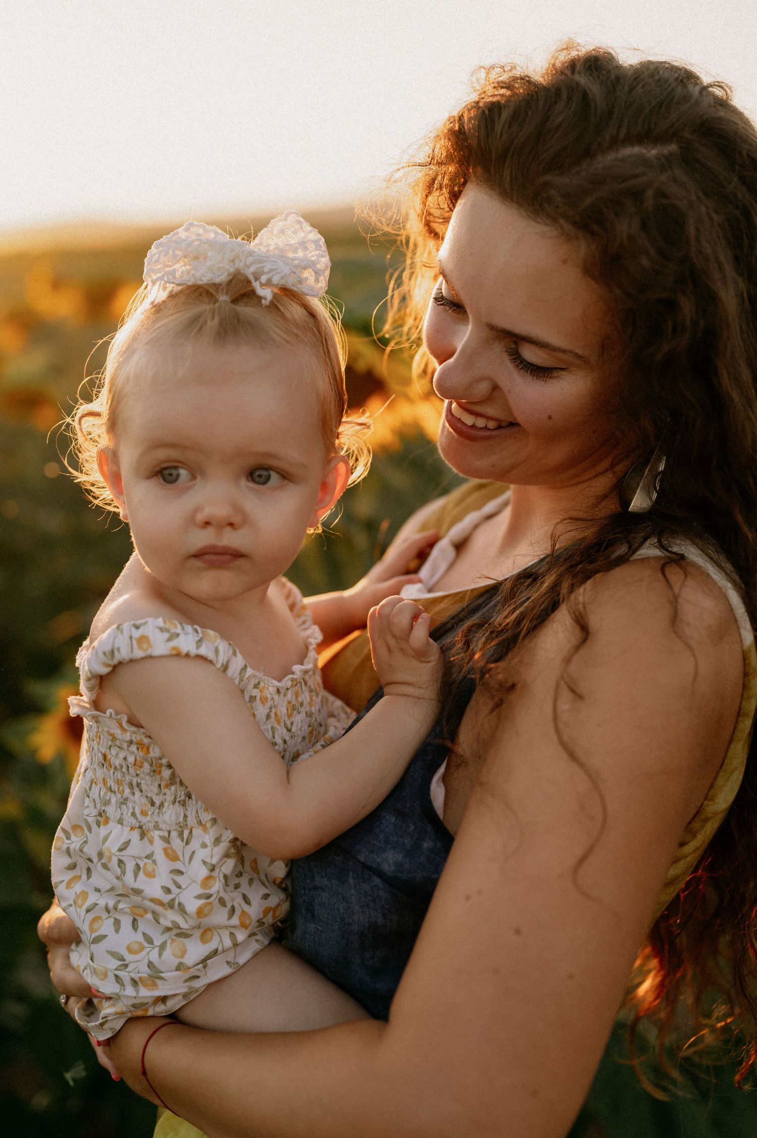 A smiling mother with curly hair holds her baby girl in a white bow headband in a sunflower field at golden hour.