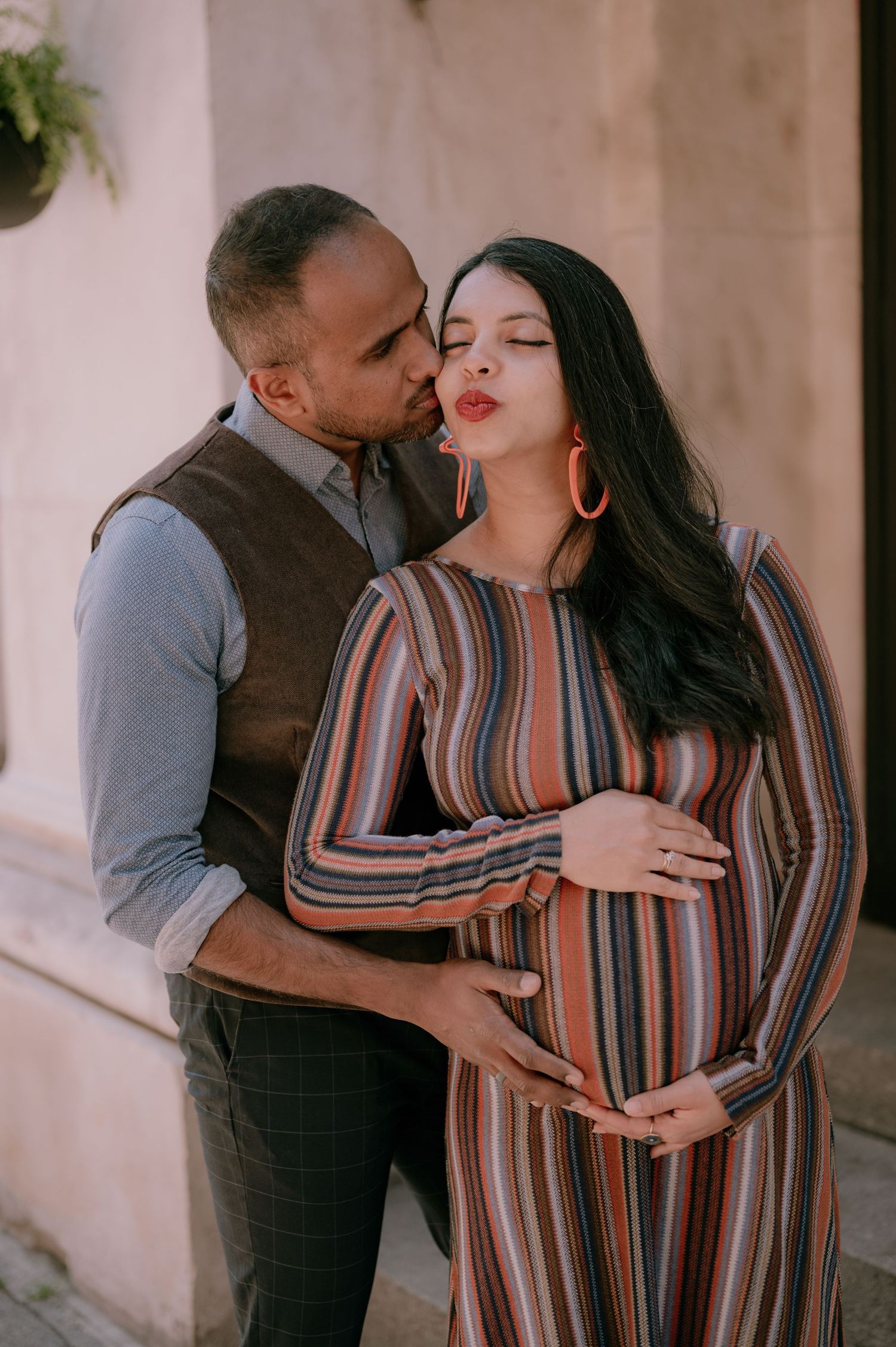 A man tenderly kisses a pregnant woman in a striped dress while cradling her baby bump during a maternity photo shoot.