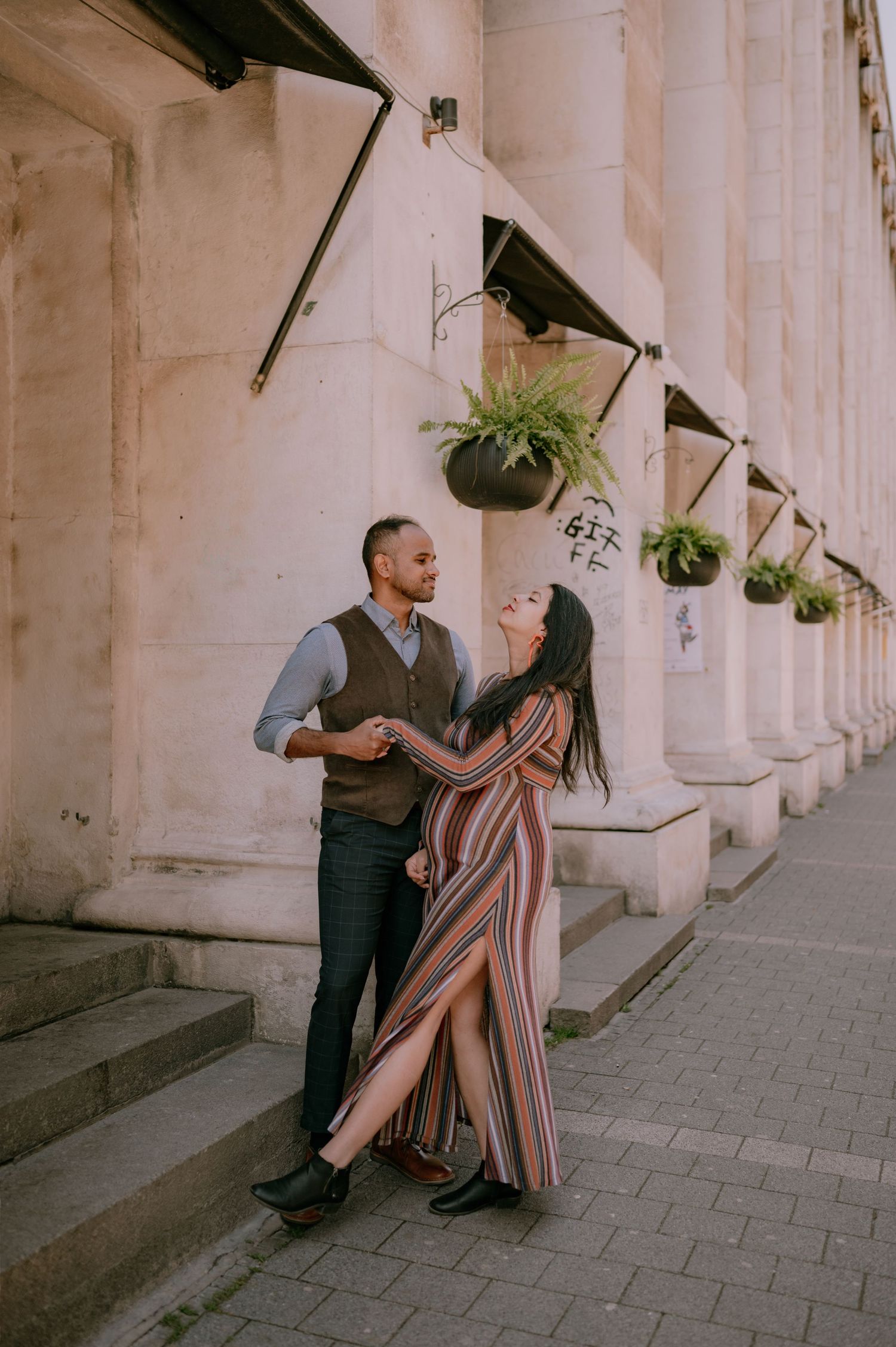 A couple shares a romantic moment outside a classical building with hanging fern planters on a cobblestone street.