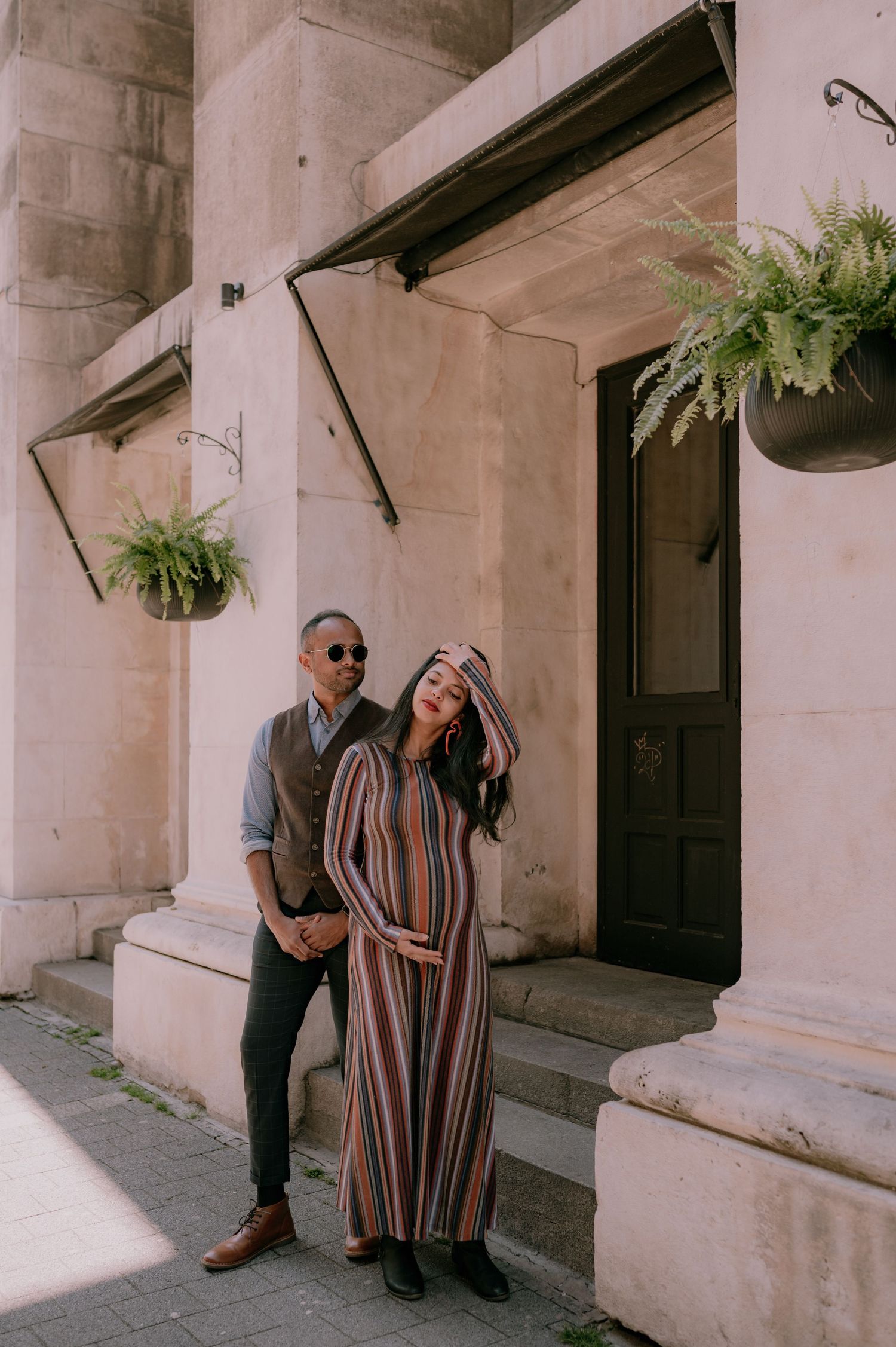 Couple posing outside a historic building, woman in striped maxi dress cradling baby bump, man in vest and sunglasses beside her.