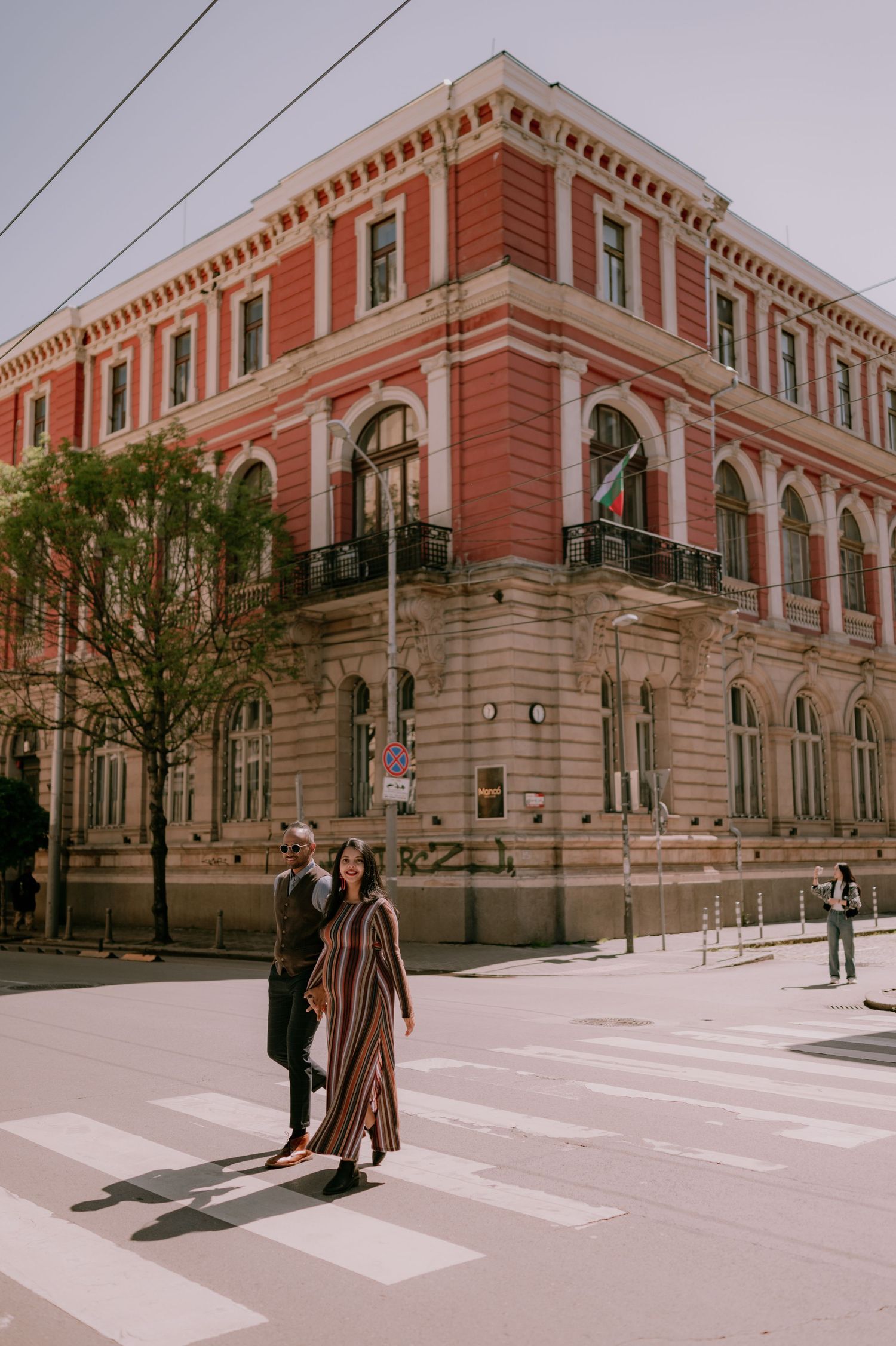 Couple crossing a zebra crossing in front of a grand red and beige historic building on a sunny day.