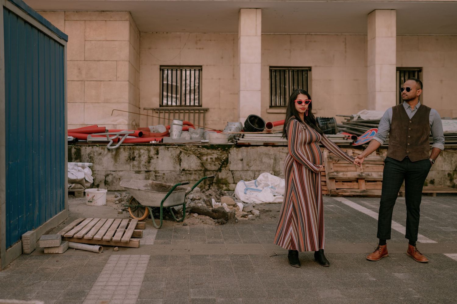A stylish couple poses in an urban setting, woman in striped maxi dress, man in vest and sunglasses.
