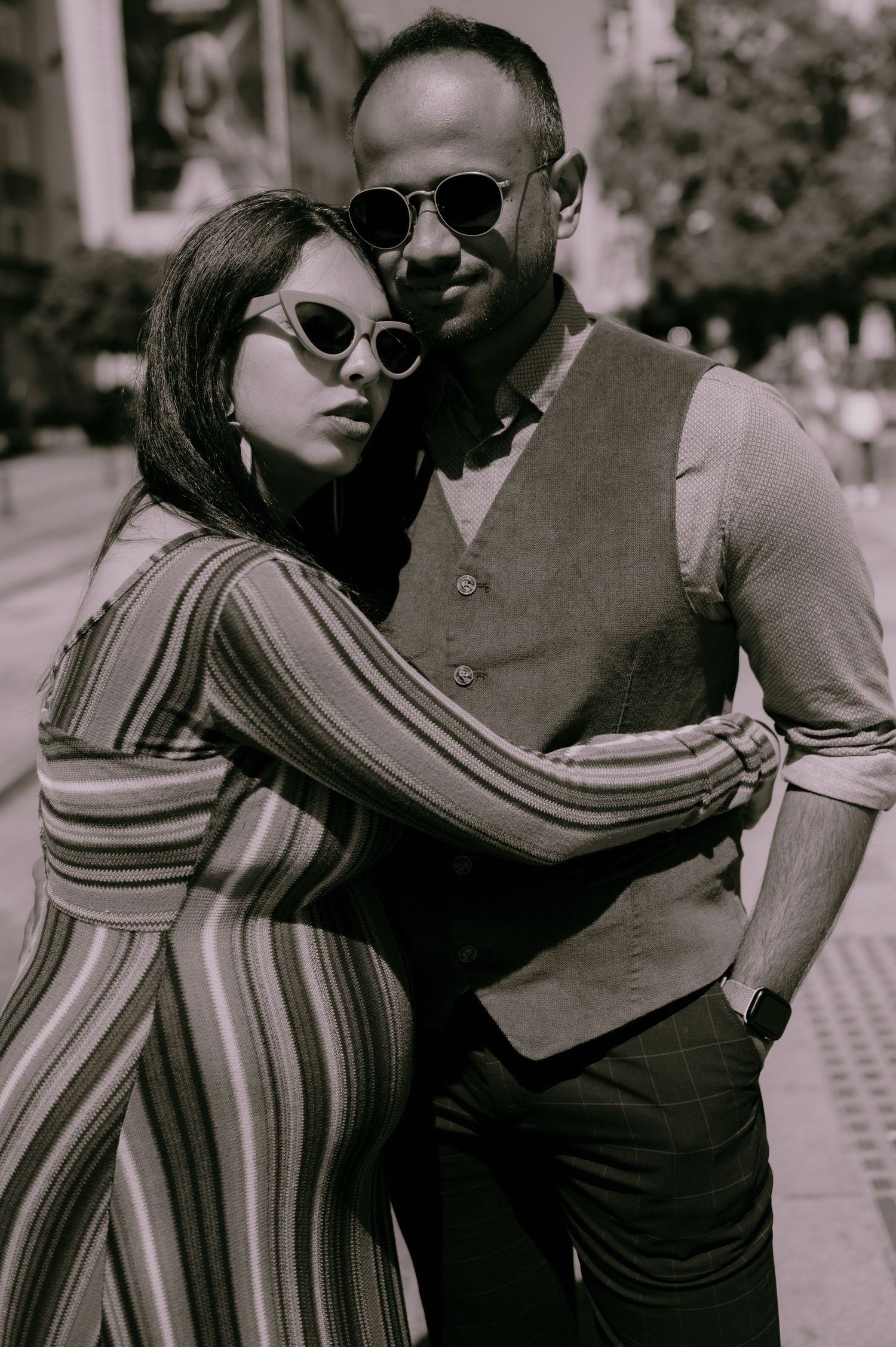 A stylish couple poses on a city street in sepia tone, wearing sunglasses and fashionable attire.