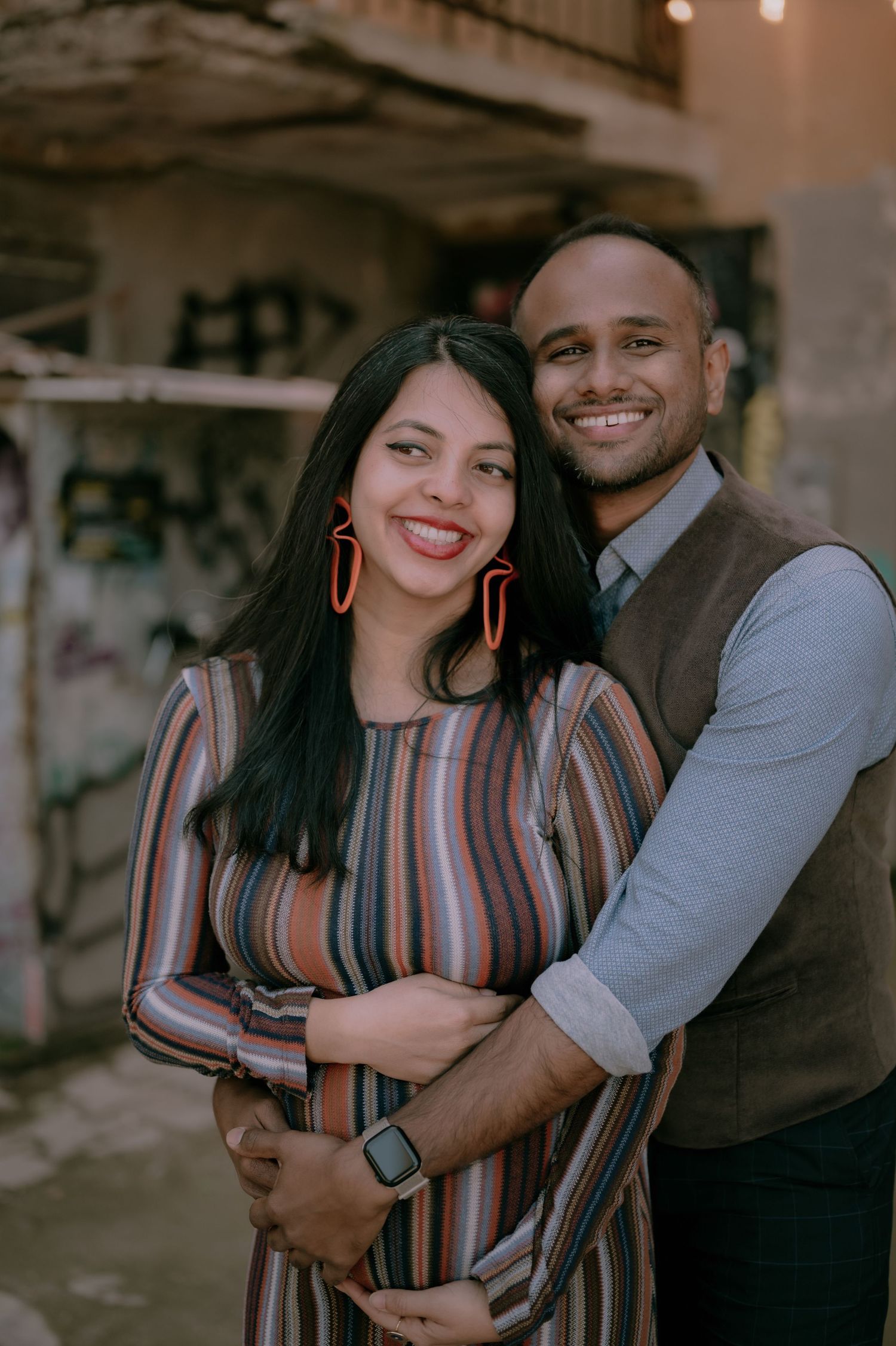 A smiling couple embraces outdoors, woman in striped dress with red earrings, man in gray vest, urban graffiti background.
