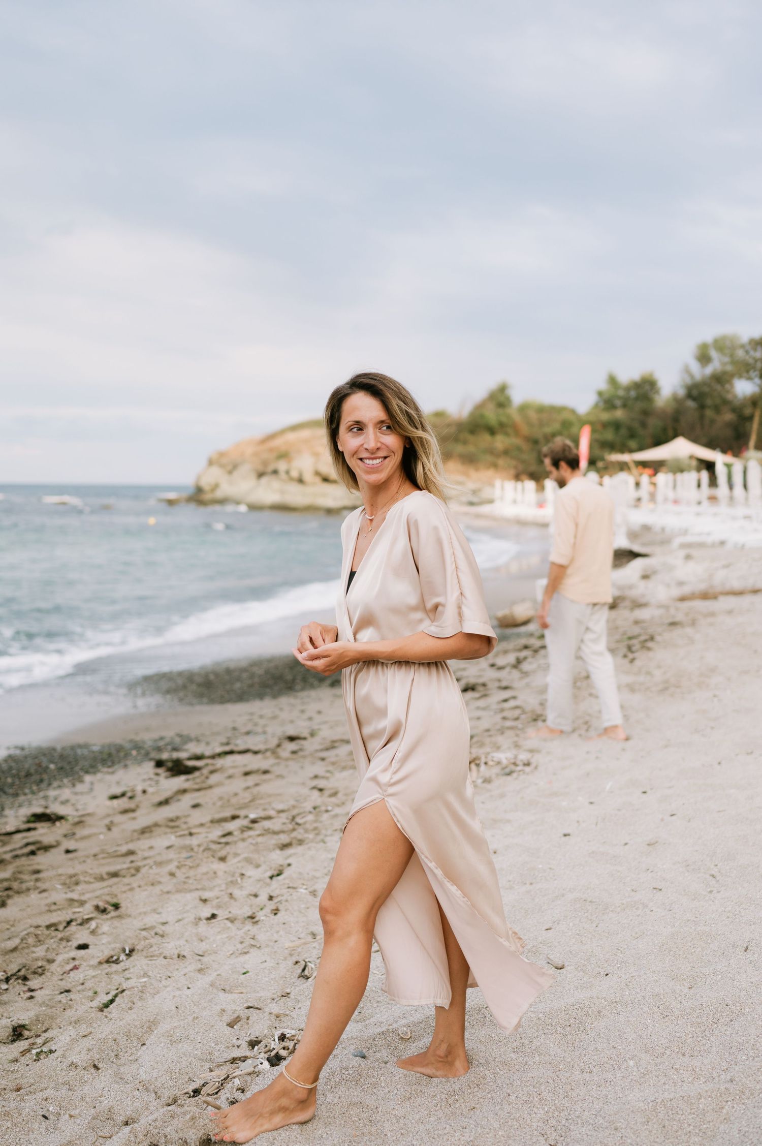 A smiling woman in a beige satin dress walks barefoot on a sandy beach with calm ocean waves behind her.