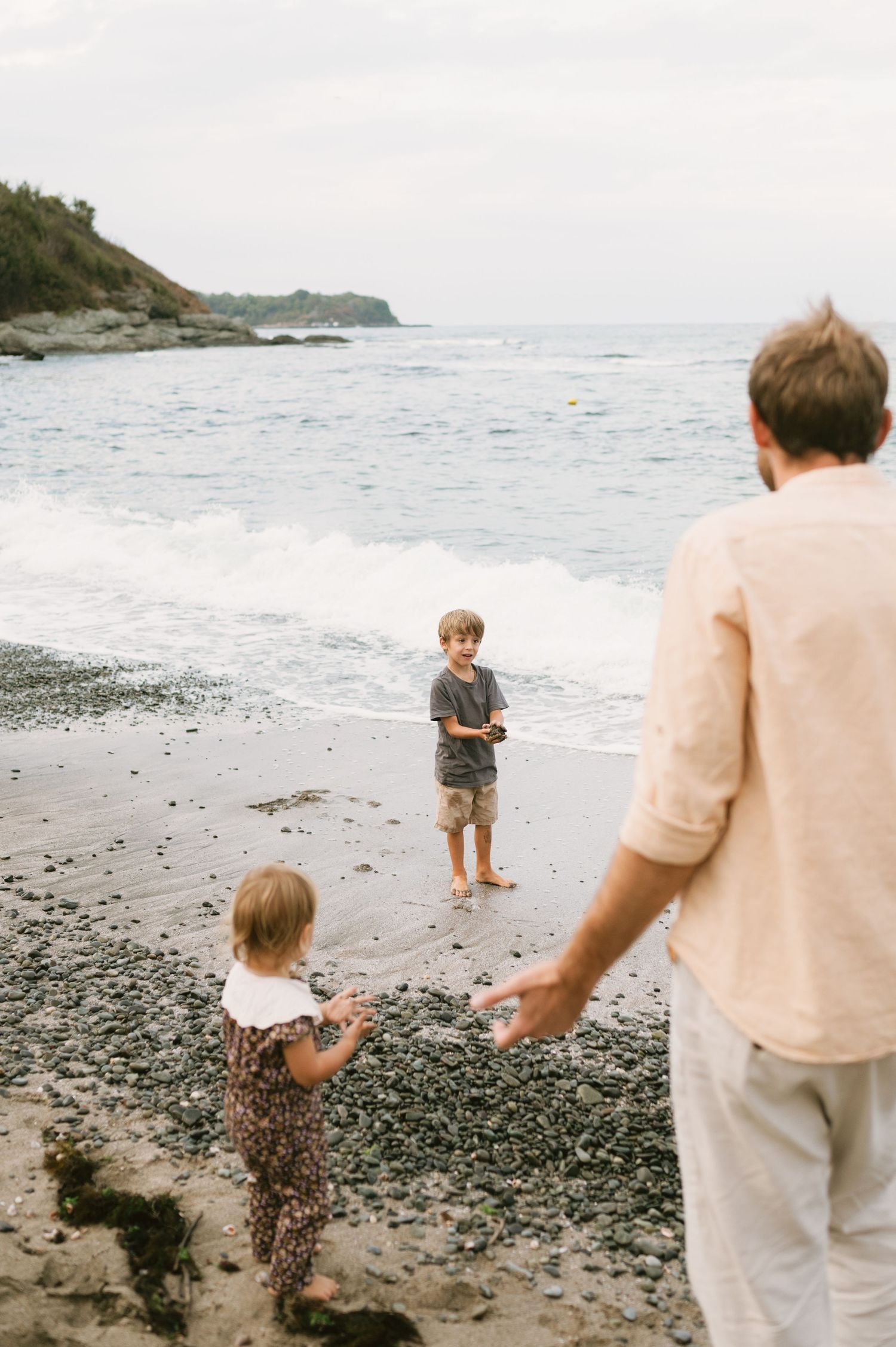 Father and two young children playing on a rocky beach with ocean waves in the background.