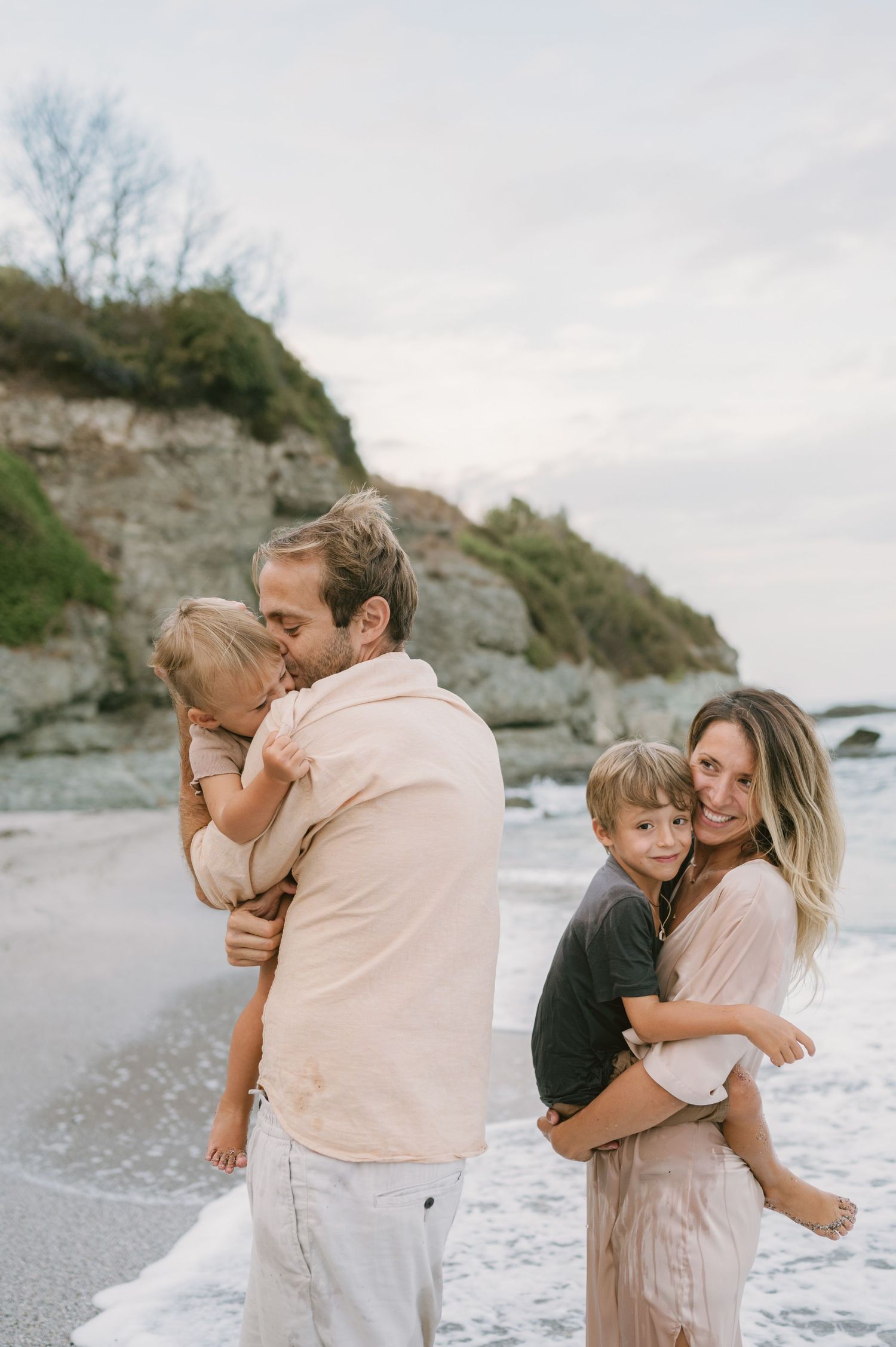 A happy family of four enjoys a beach day, with parents holding their young children near ocean waves.