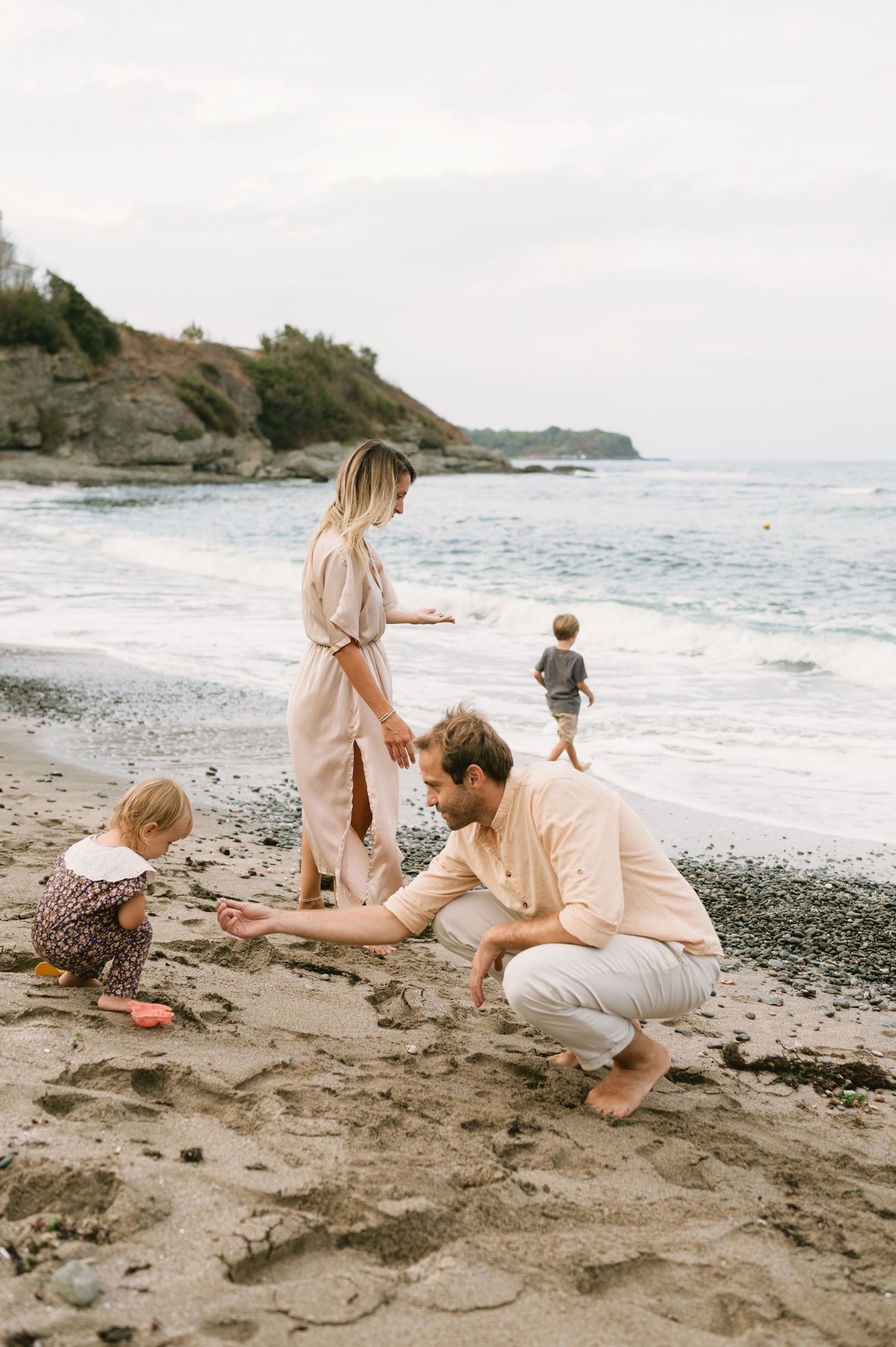 Family playing on a sandy beach, parents and two young children enjoying the ocean shoreline on a cloudy day.