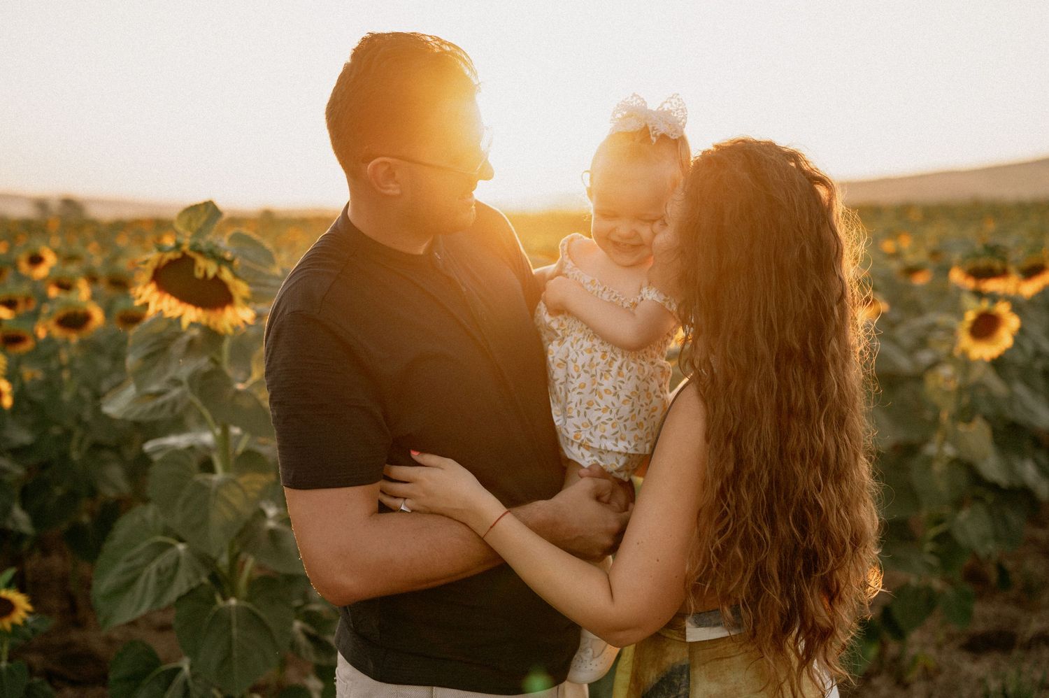 Couple holding toddler girl in sunflower field at golden sunset hour.