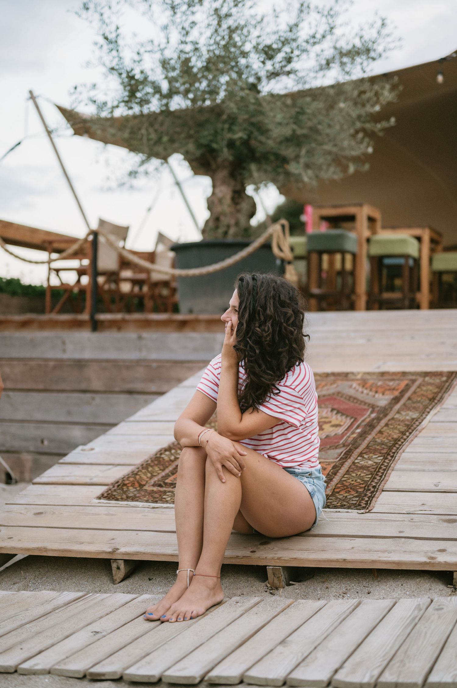Young woman in striped top and denim shorts sitting on wooden deck near a rustic outdoor lounge area.