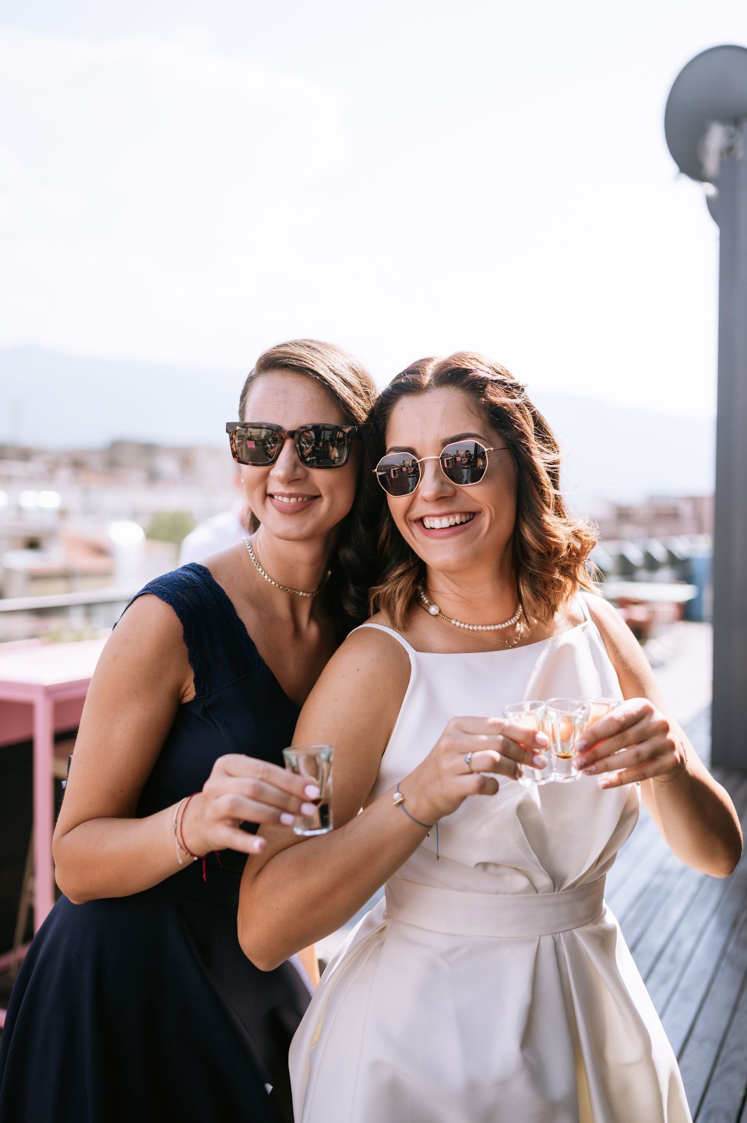 Two friends in black and white dresses share drinks and smiles on an outdoor terrace overlooking a city view.