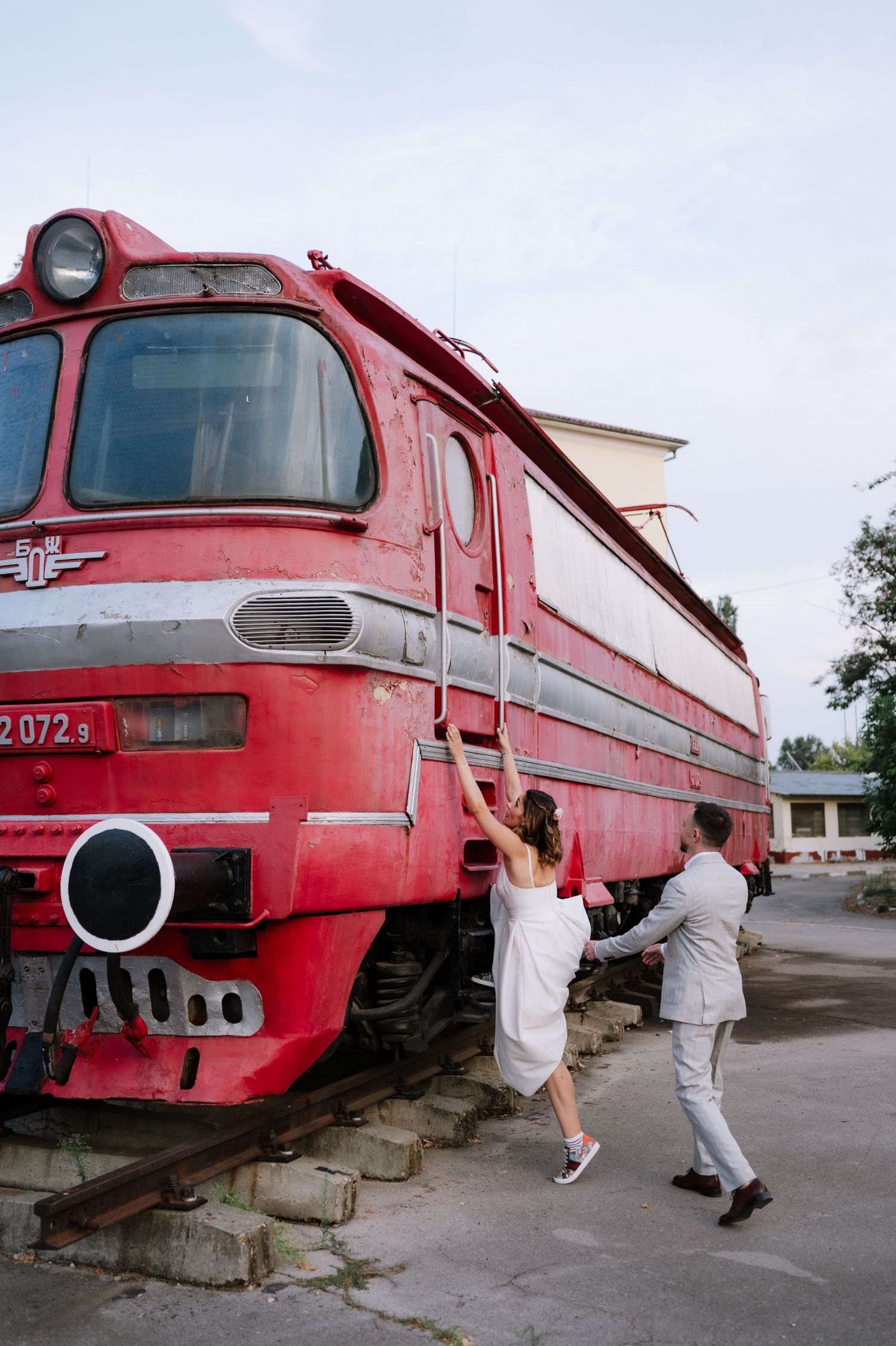 Two people playfully jump and pose in front of a vintage red locomotive train at a railway station.