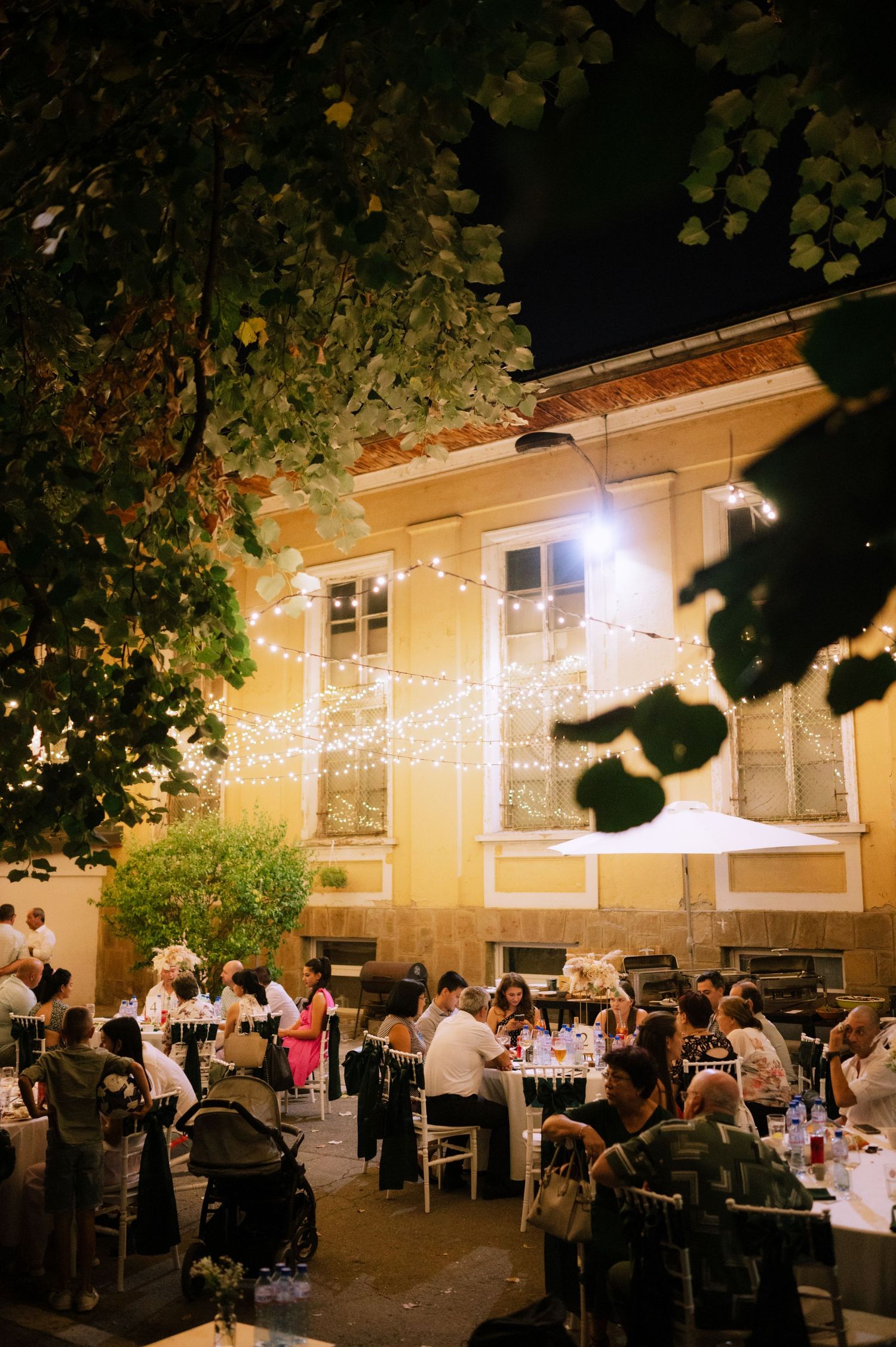 String lights illuminate an outdoor evening wedding reception with tables and guests dining under a warm glow.